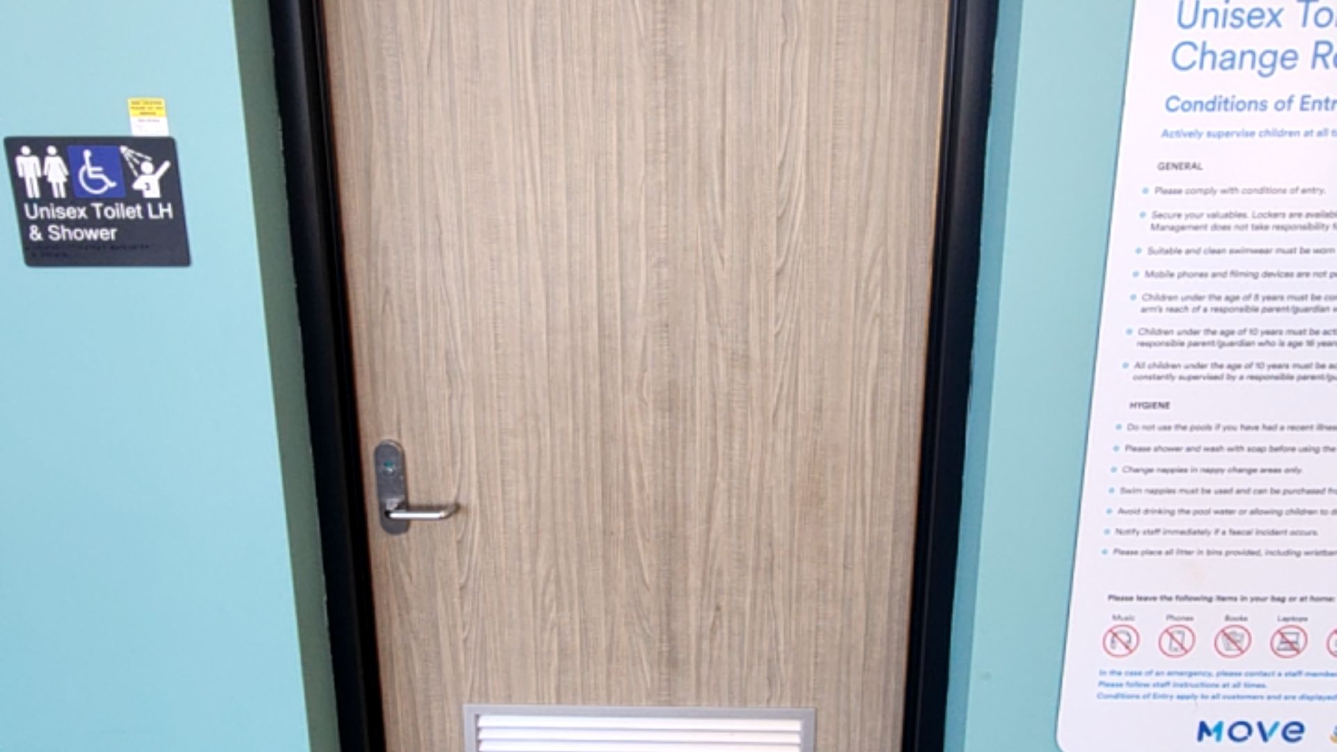 Light timber door with silver lever handle and indicator lock. To the left, a black sign with white icons and braille for Unisex Toilet LH and Shower. Light blue walls.