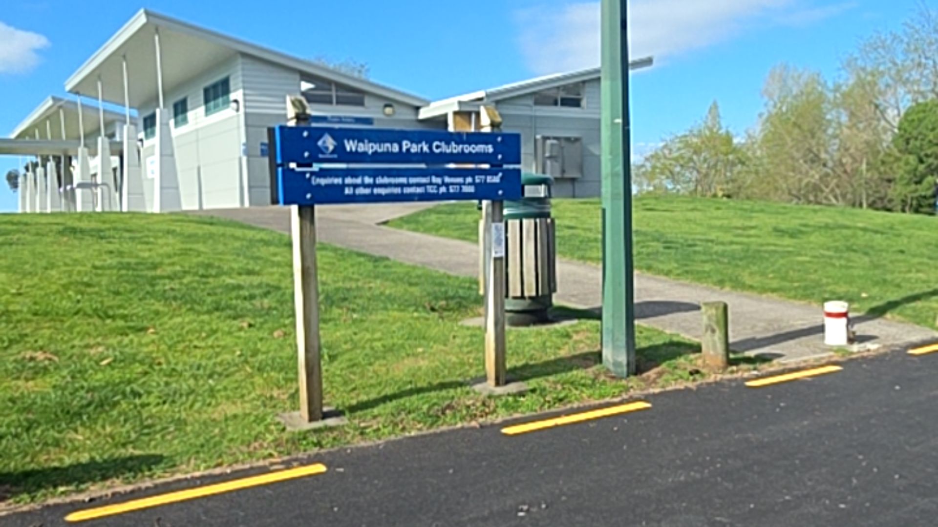 Blue Waipuna Park Clubrooms sign beside a concrete pathway with a gentle uphill gradient. The path leads from a black asphalt road with yellow markings to a modern grey building.