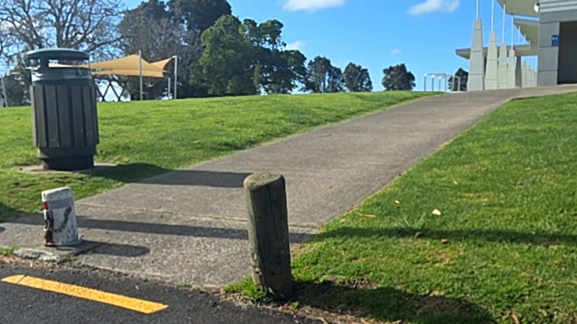 Concrete pathway with an upward gradient. A wooden bollard and short white post mark the entrance. A green rubbish bin sits on the grass to the left.