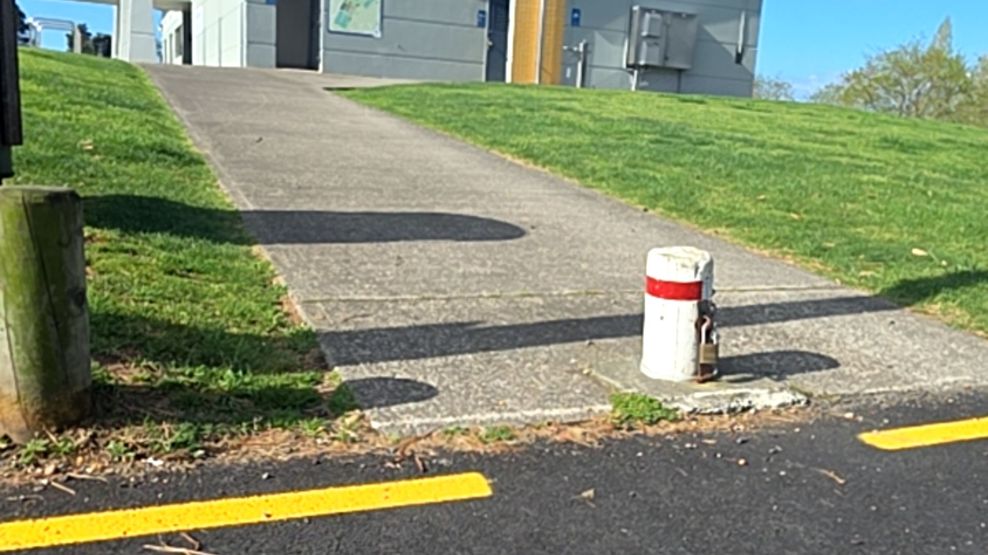 Sloping concrete pathway leads from an asphalt road with a yellow line. A white and red bollard sits on the right side of the path entrance. Grassy slopes border the path on both sides.