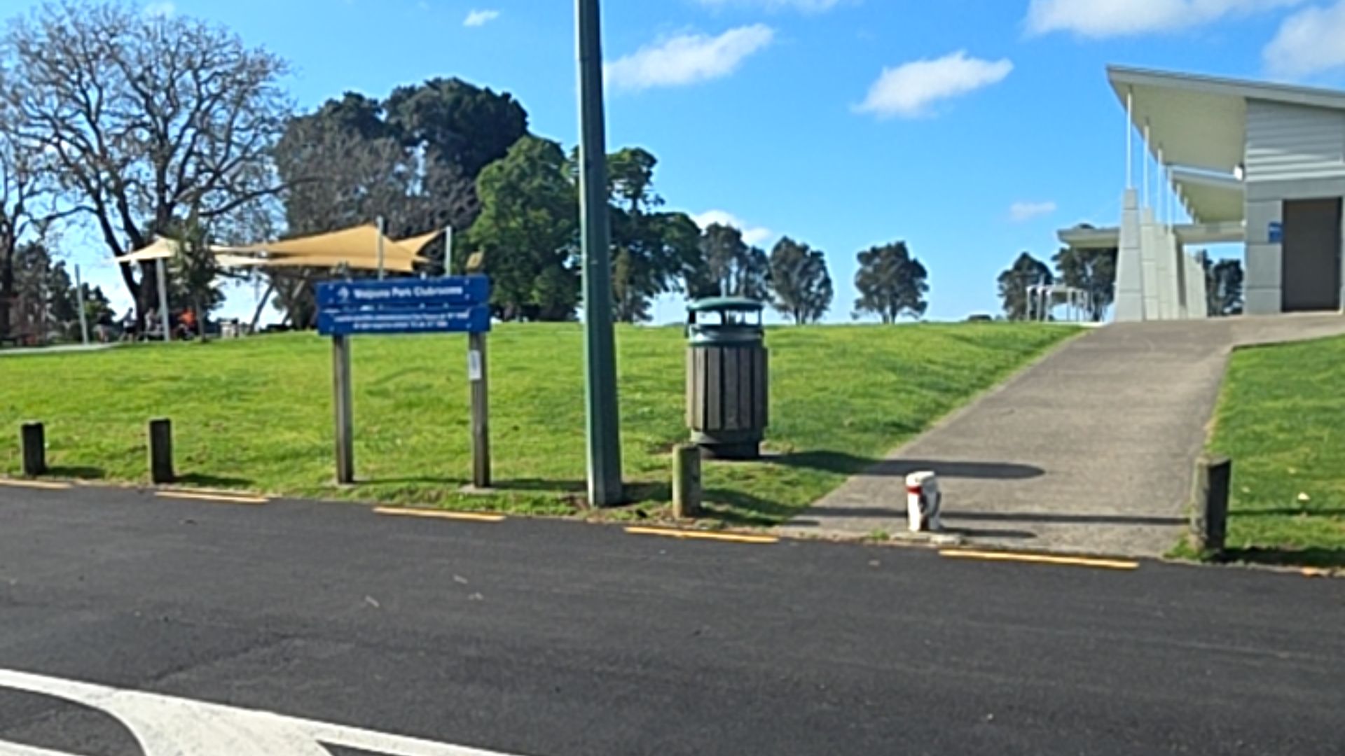 Wide sloping concrete pathway leads from an asphalt road past a blue sign and timber-clad rubbish bin to a white building. Wooden bollards line the grassy verge.