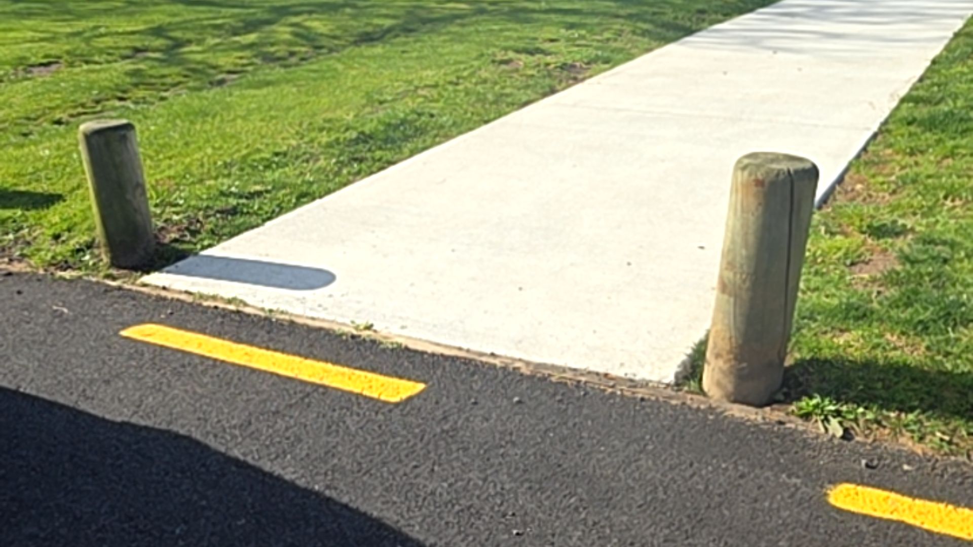 Level concrete pathway joins a bitumen surface with yellow line markings. Two weathered timber bollards flank the entrance. Firm, even surfaces throughout.