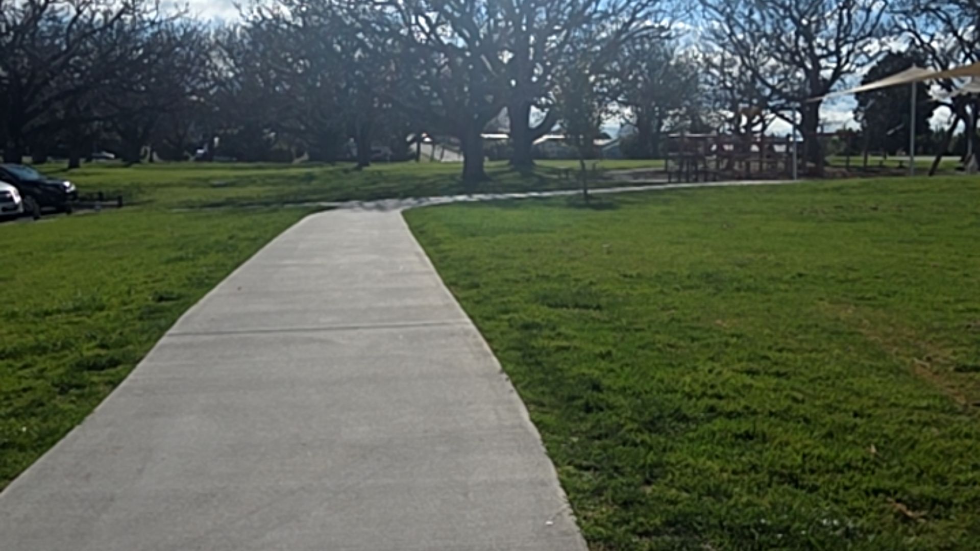 Wide, level grey concrete pathway leading through a grassy park toward large bare trees and a timber playground with a shade sail. The path is smooth and clear of obstructions.