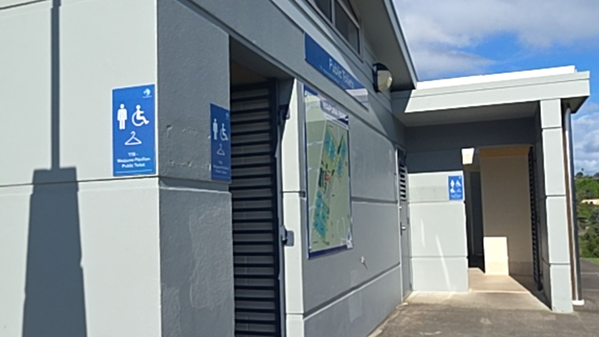 Level concrete approach to a grey public toilet block. Blue wall signage displays male, wheelchair, and coat hanger symbols next to a dark slatted doorway and an open alcove entrance.