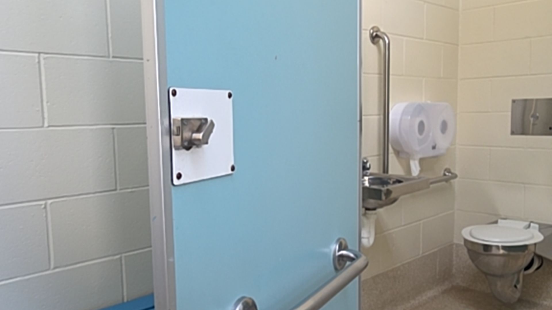 Blue door with silver latch and horizontal grab rail. Inside, a stainless steel toilet, sink, and multiple grab bars mounted on light-coloured tiled walls over a level floor.