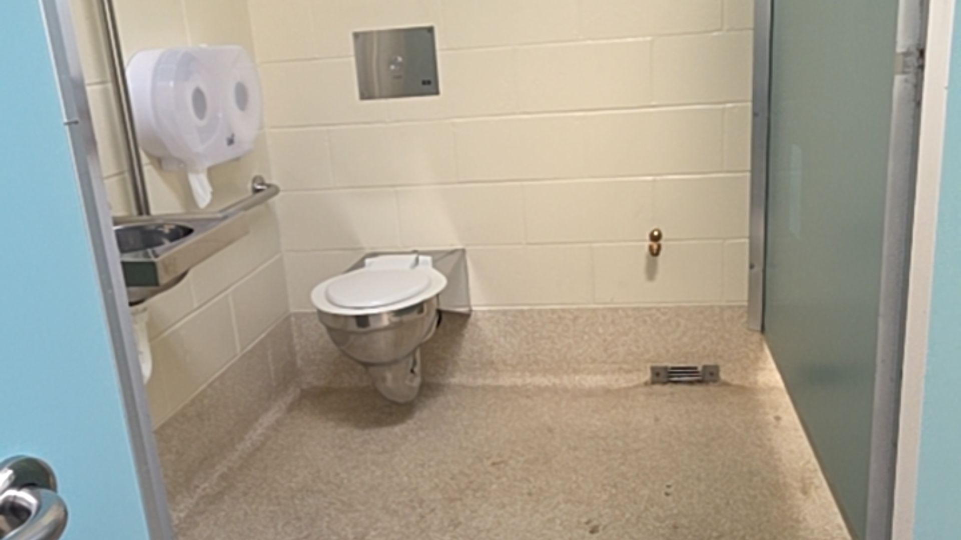 Spacious toilet with stainless steel pan and basin, horizontal grab rail, and level slip-resistant floor. Cream block walls with ample room for manoeuvring and wide doorway.