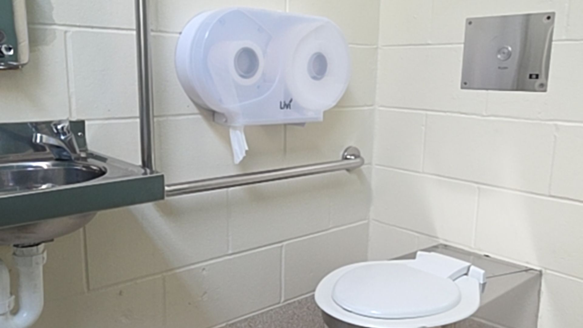 Stainless steel toilet with white seat and horizontal grab bar. Translucent toilet paper dispenser and push-button flush panel on off-white brick walls. Stainless steel basin to the left.