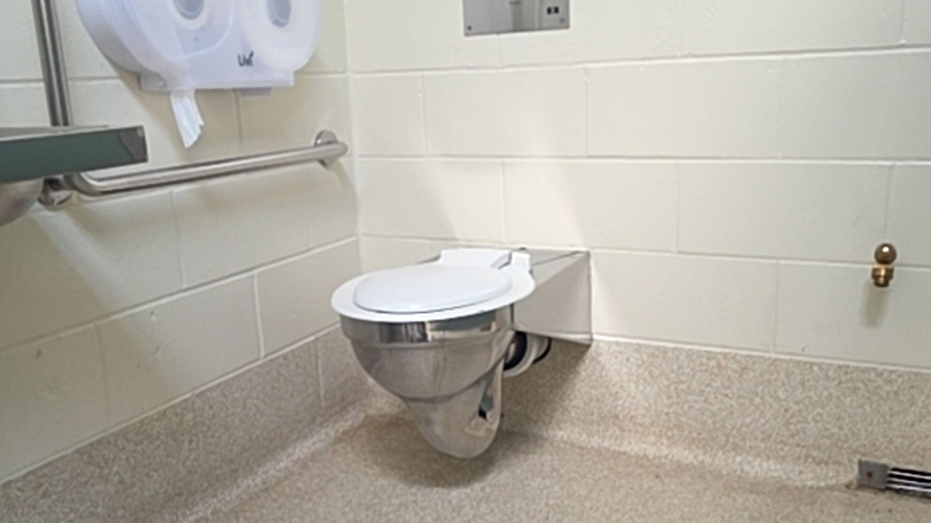 Wall-mounted stainless steel toilet with white lid. Horizontal grab bar on off-white tiled wall to the left. Level, slip-resistant neutral flecked flooring with clear space.