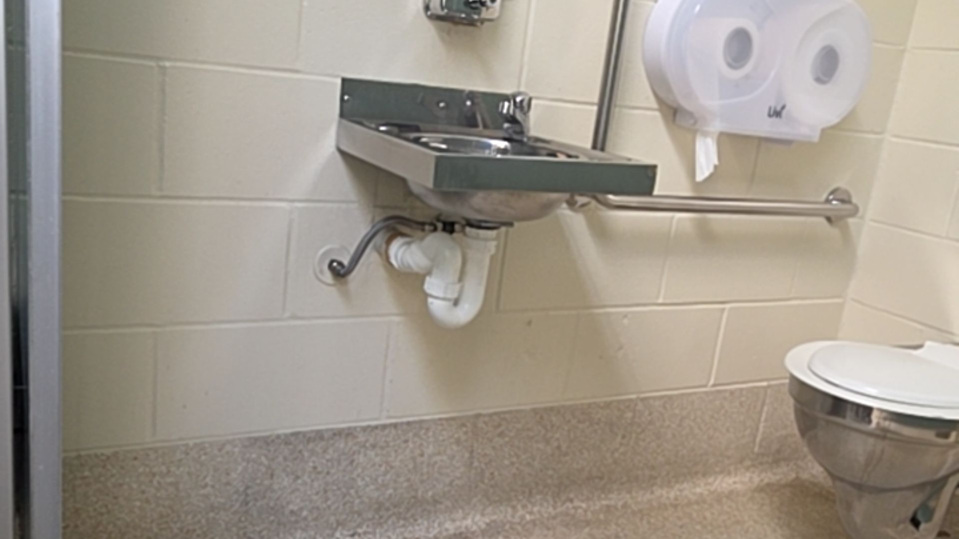 Wall-mounted stainless steel sink with knee clearance and exposed white plumbing. Stainless steel toilet with white lid. Horizontal grab bars on light-coloured block walls. Speckled floor.