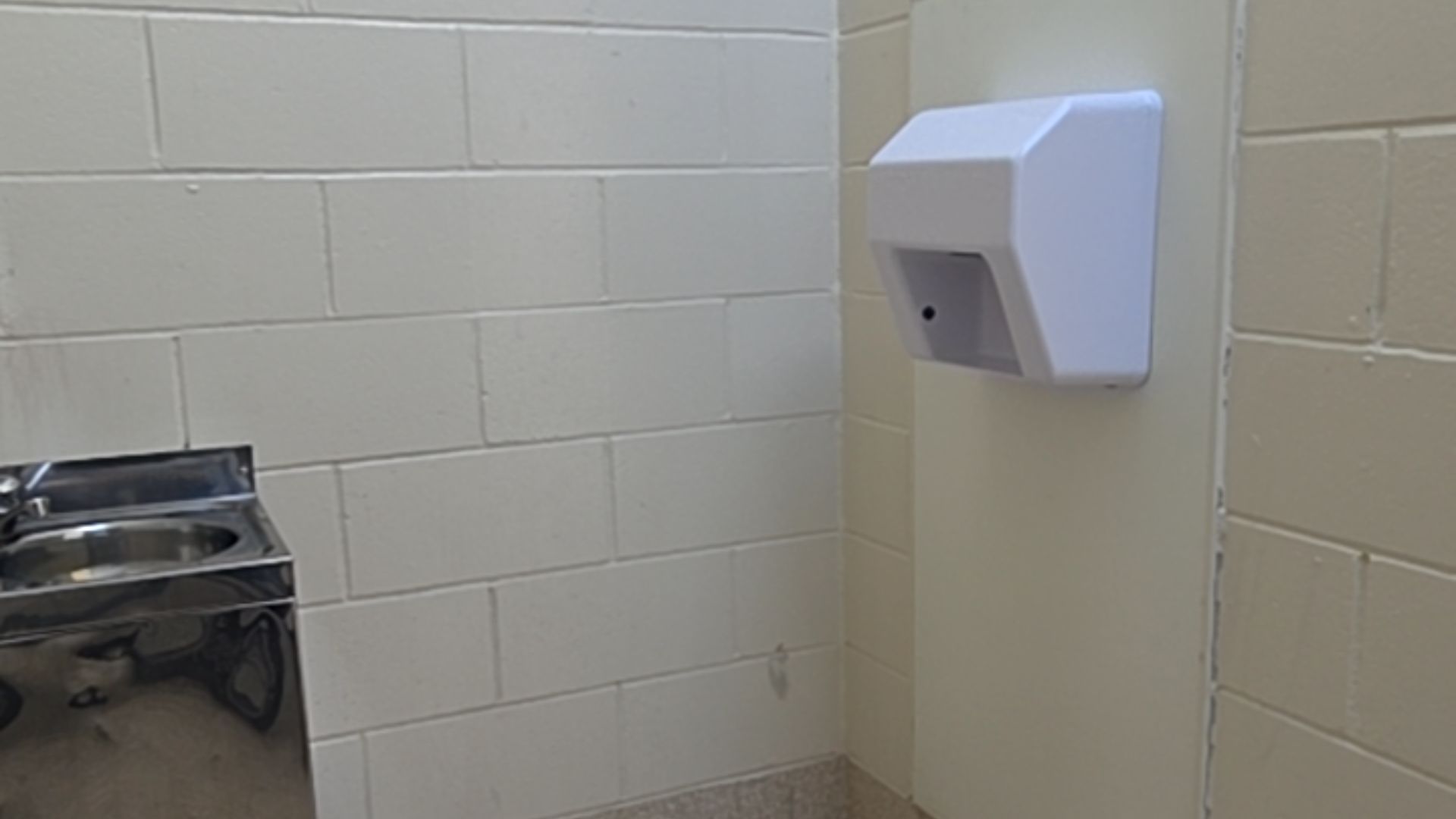 Stainless steel basin and white wall-mounted hand dryer. Walls are light-coloured cinder blocks, and the floor is a level, speckled surface.