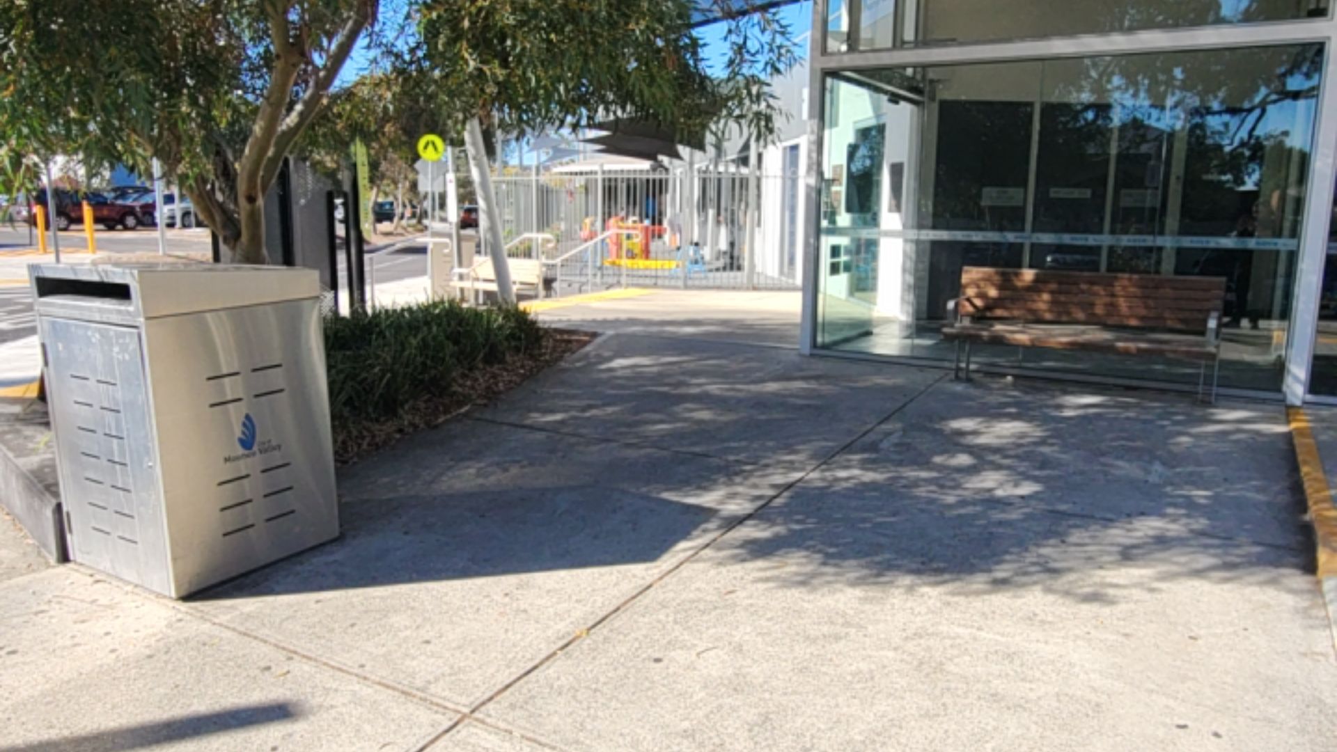 Level concrete path leads to a wide glass entrance. A timber and metal bench is on the right. A silver rubbish bin and garden bed are on the left. Pedestrian crossing sign in background.