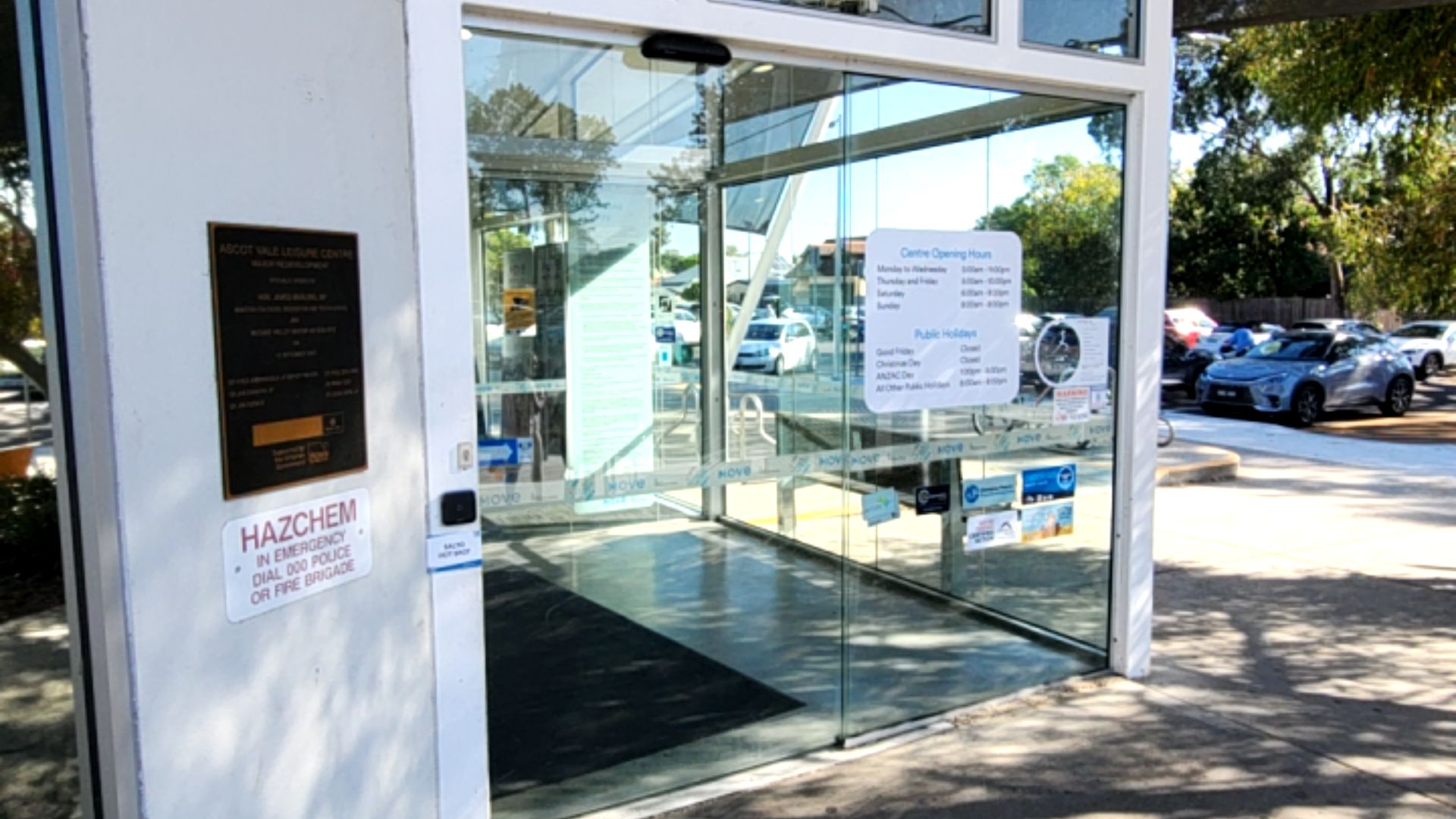 Level concrete entrance with automatic glass sliding doors. Signage includes opening hours and a Hazchem warning. Flat, unobstructed indoor and outdoor floor surfaces.