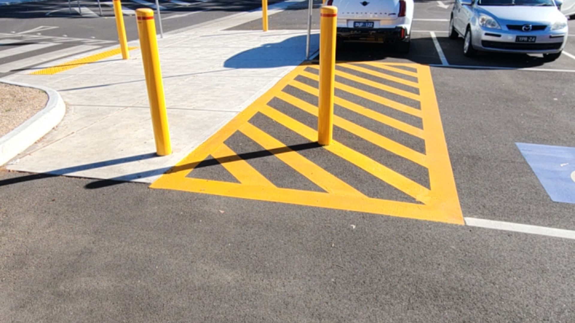 Concrete ramp with yellow tactile indicators meets a yellow hatched access zone. A tall yellow bollard is positioned within the hatched markings next to an accessible parking space.
