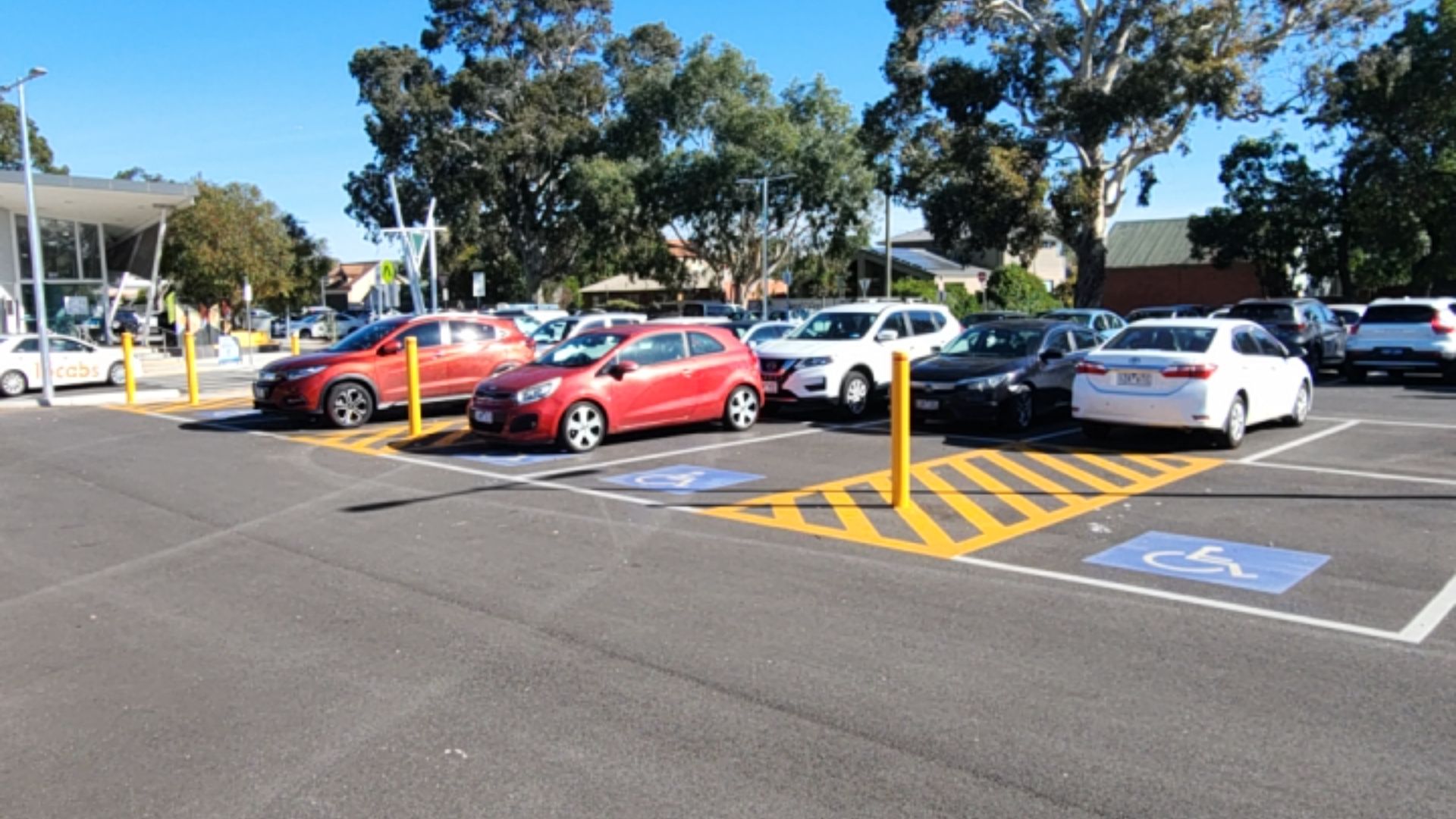 Four accessible parking spaces with blue wheelchair symbols and yellow hatched shared zones on level asphalt. Yellow bollards are located at the front of the shared zones.