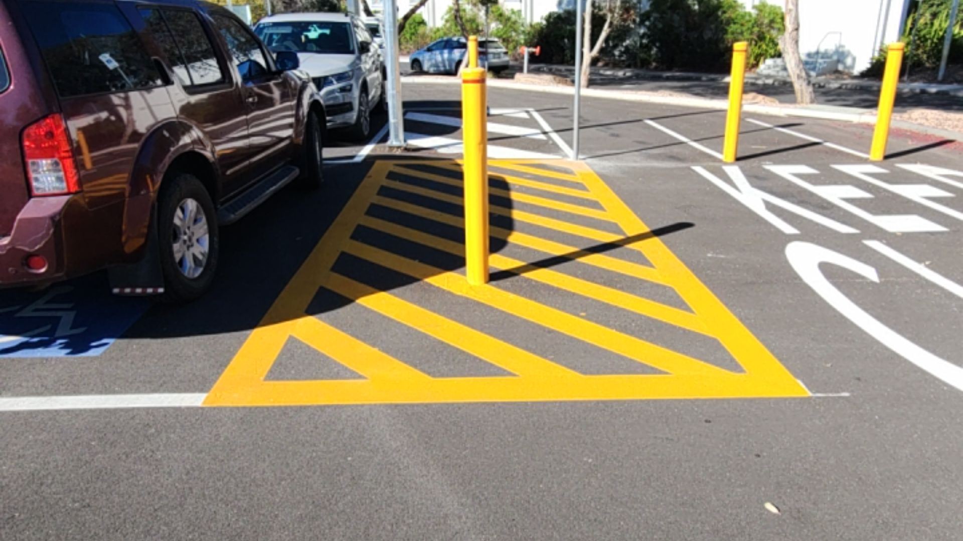 Maroon SUV in a parking space with blue wheelchair symbol next to a yellow hatched zone. A yellow bollard stands in the centre. White "KEEP CLEAR" text is painted on the level bitumen ground.