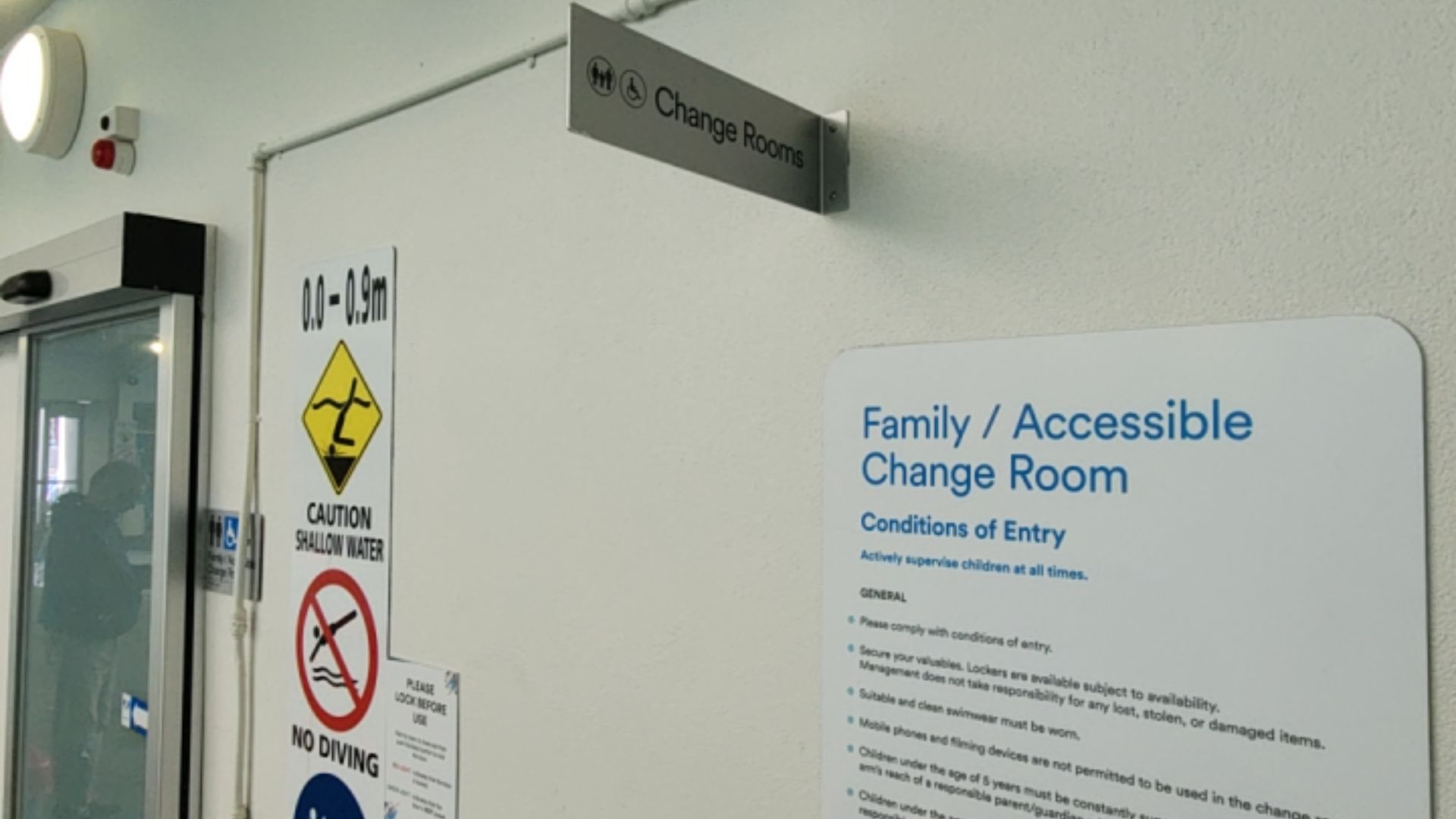 Signage for family and accessible change rooms on a white wall. A silver sign features a wheelchair icon. A large white board lists entry conditions next to pool safety signs and a glass doorway.