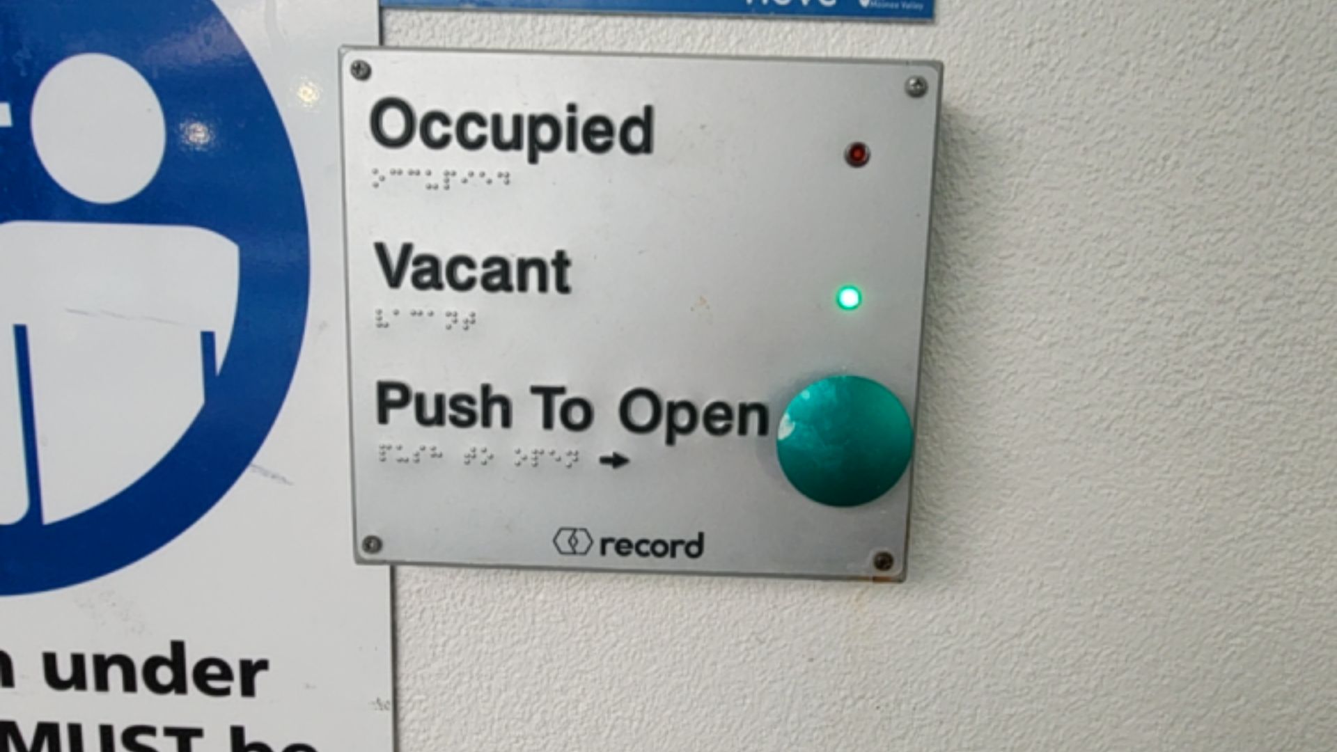 Silver control panel with 'Occupied', 'Vacant', and 'Push To Open' text and Braille. Green light lit next to 'Vacant' above a large green push button. Partial blue accessibility symbol on left.