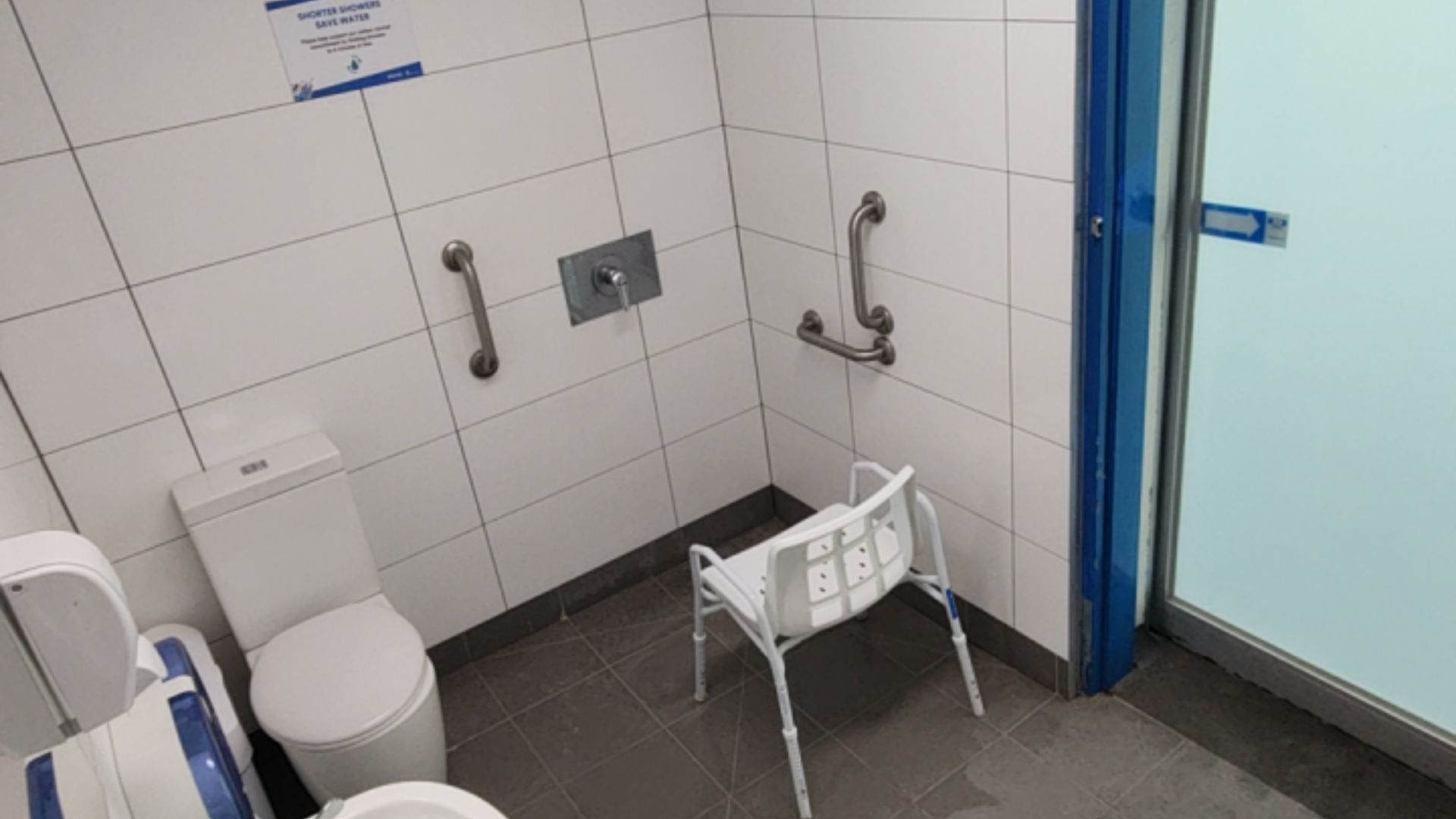 Level-access bathroom with dark grey tiles, white toilet, adjustable shower chair, silver grab bars on white tiled walls, and a blue-framed frosted glass sliding door.