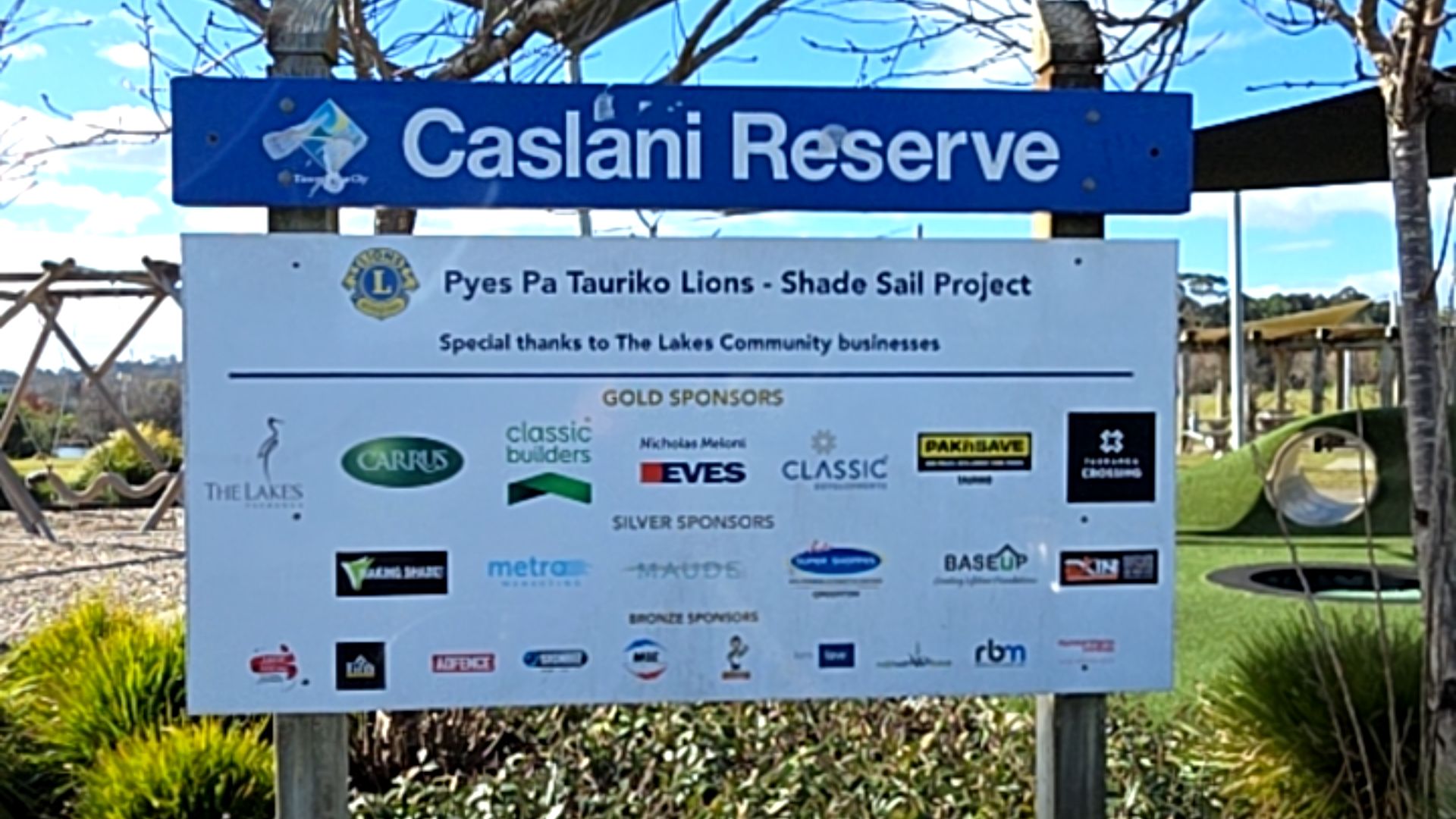 Caslani Reserve signage on timber posts. Lower white board lists project sponsors. In the background, a grassed park includes a concrete tunnel and wooden playground structures.