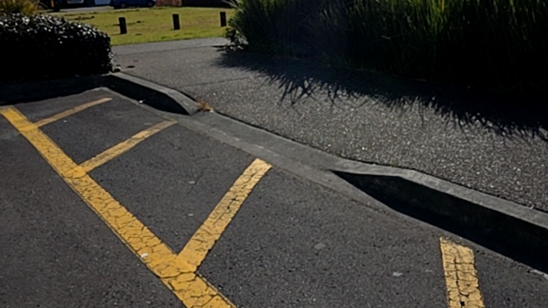 Asphalt parking space with yellow diagonal line markings next to a concrete kerb ramp. The ramp leads to a level, textured bitumen pathway.