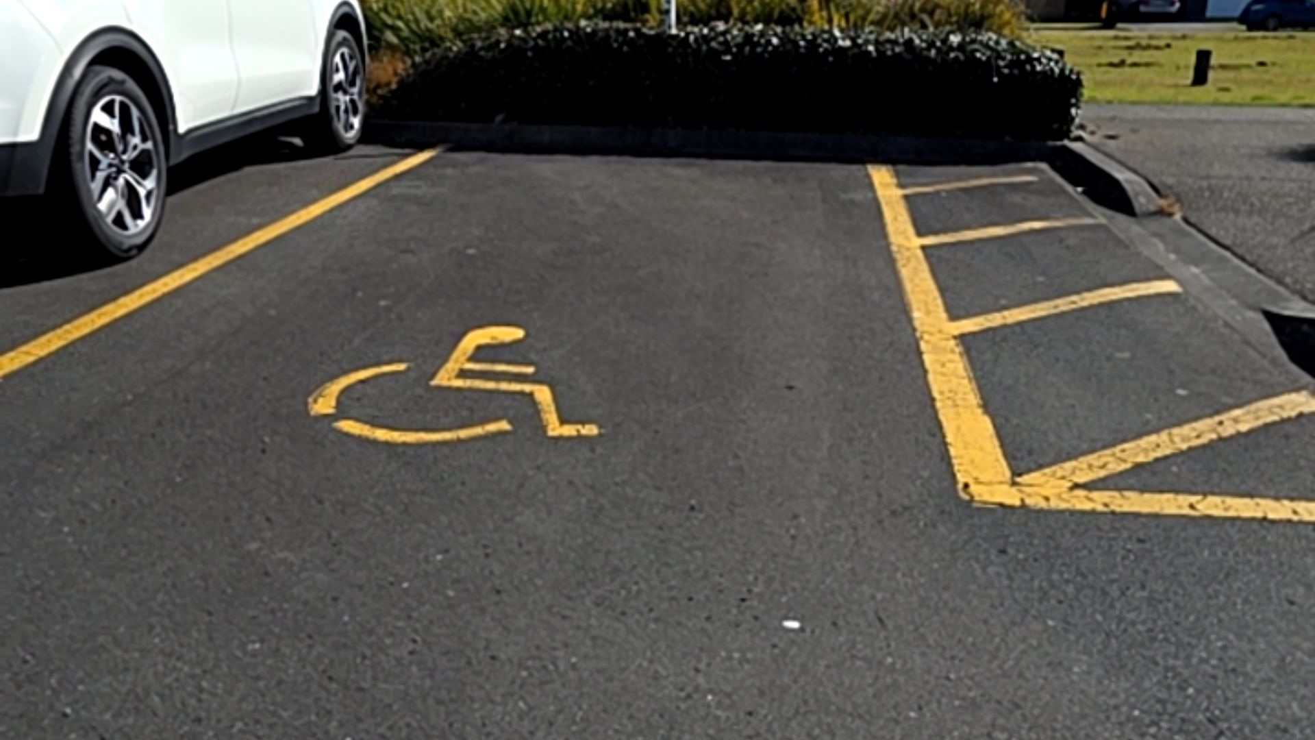 Level asphalt accessible parking space marked with a yellow wheelchair symbol and a yellow-hatched shared zone. A white car is parked in the adjacent standard space.