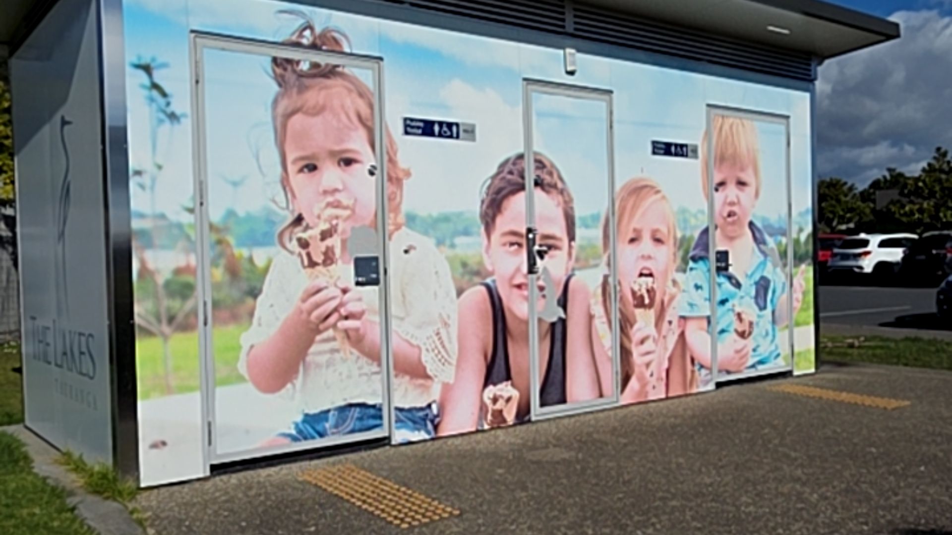 Level asphalt pathway with yellow tactile ground surface indicators leading to public toilet doors. Mural-covered building with wheelchair accessibility symbols on door signage.