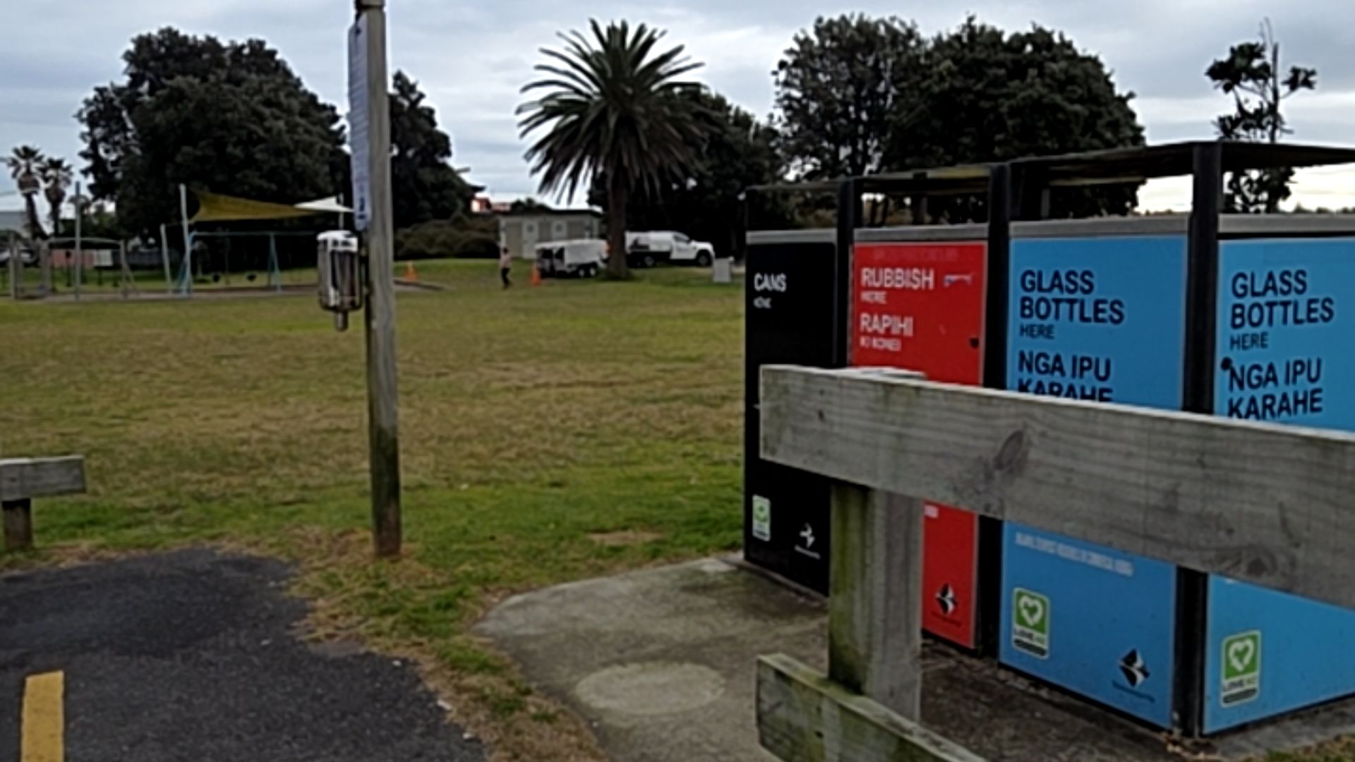Colour-coded waste bins for cans, rubbish, and glass on a level concrete pad, with English and Māori signage. Adjacent to an asphalt path, wooden fence, and grassy park area.