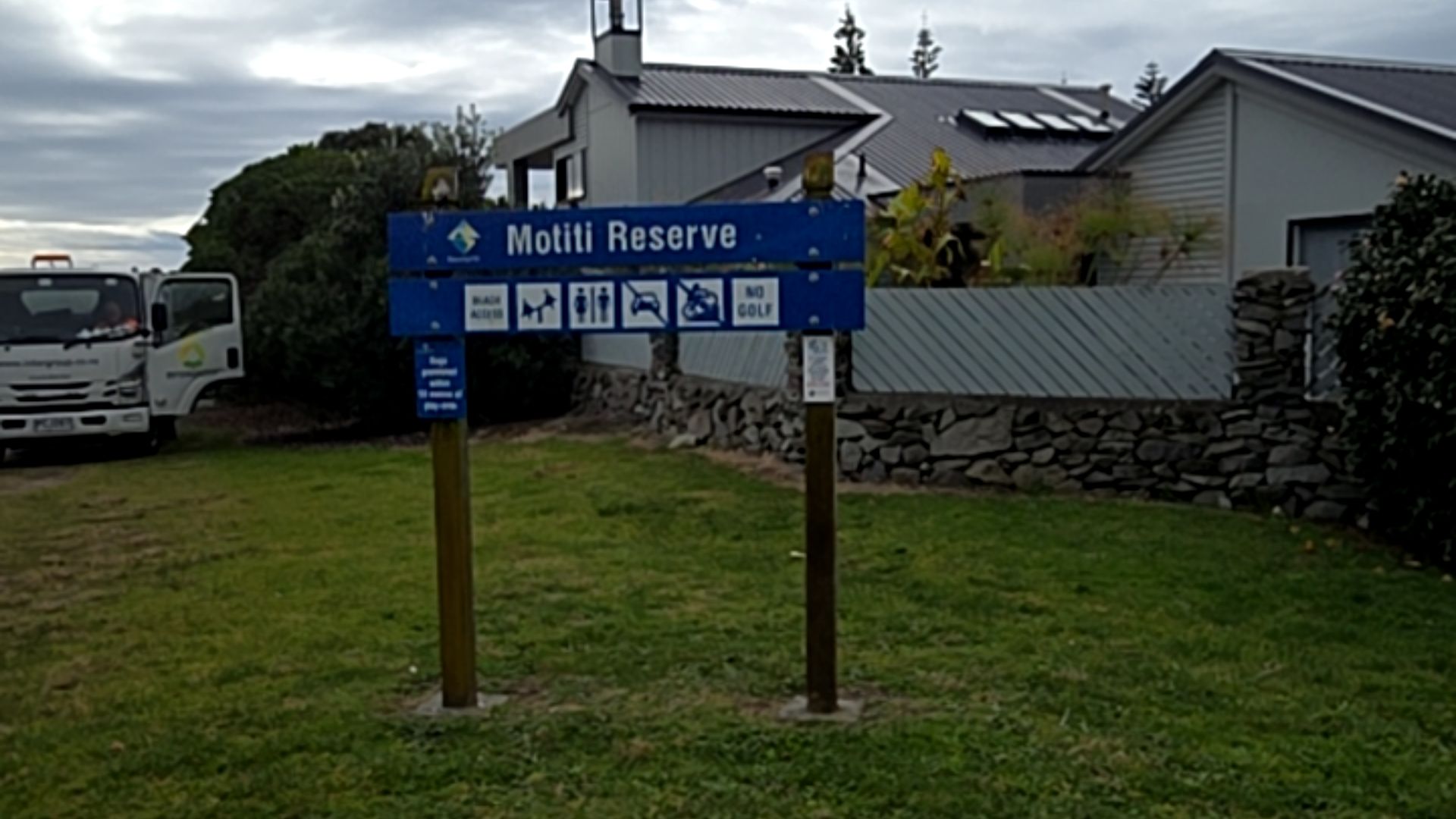 Blue timber Motiti Reserve sign on grass. Icons show beach access, toilets, dog rules, and no golf. A grey house with a stone wall and a white truck are in the background.