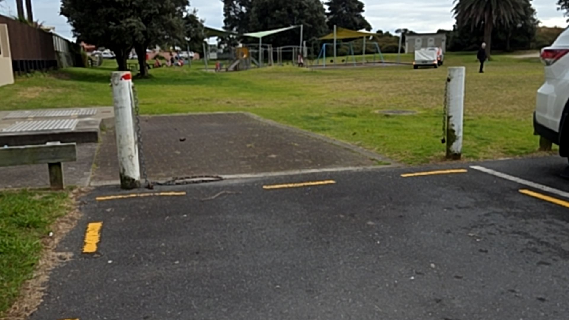 Level transition from an asphalt car park to a paved area between two white bollards. A chain lies on the ground between them. A grassy park and playground are in the background.