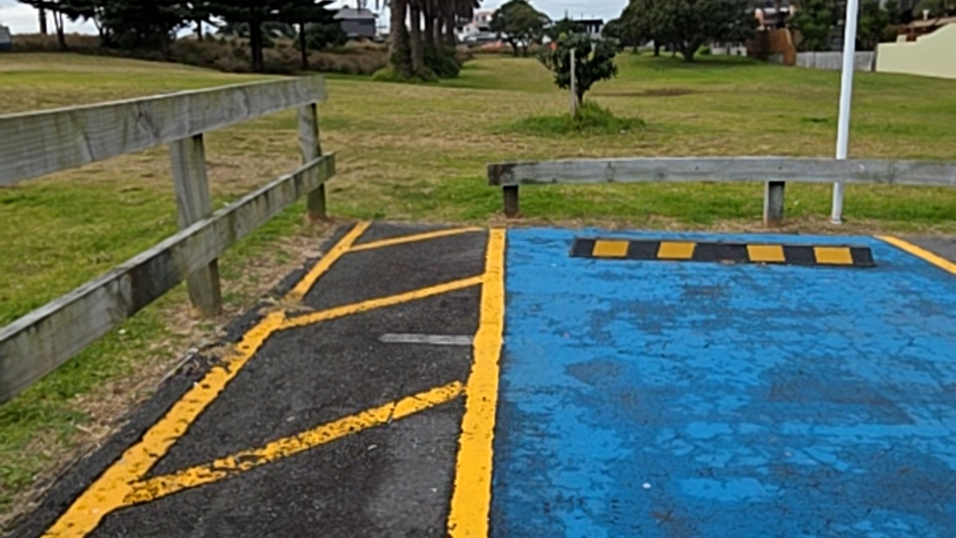 Level blue-painted parking bay with a yellow-striped access aisle. A black and yellow wheel stop marks the end. Low wooden rail fencing borders the asphalt area next to a grassy parkland.