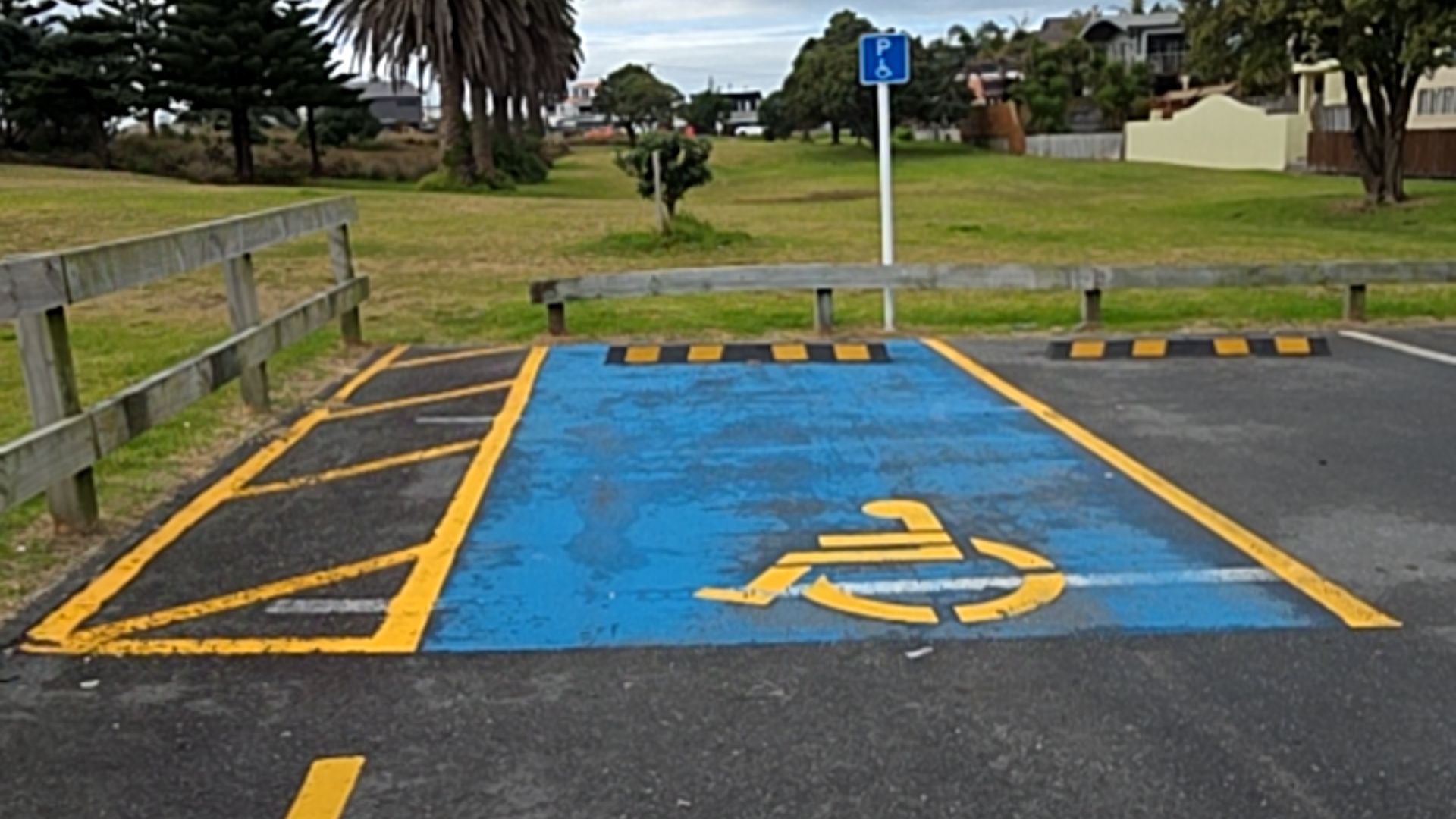 Blue accessible parking bay with yellow wheelchair symbol and yellow-hatched access zone. Blue wheelchair sign on a white pole at rear. Level asphalt surface with a low wooden barrier.