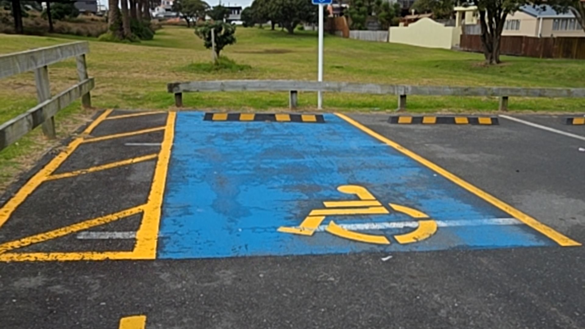 Level asphalt accessible parking space painted blue with a yellow wheelchair symbol. Includes a yellow hatched access aisle on the left and a yellow-striped wheel stop at the head.