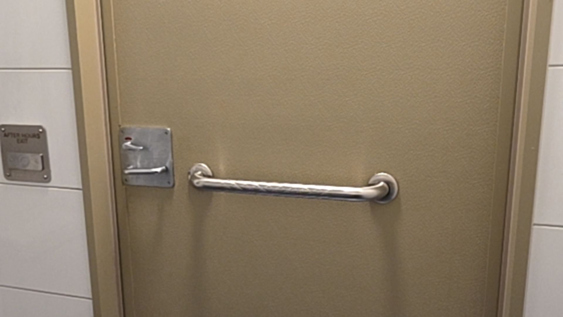 Tan door with a horizontal stainless steel grab bar. A metal plate on the left features two lever latches. Adjacent white tiled wall has an after-hours exit sign and button.