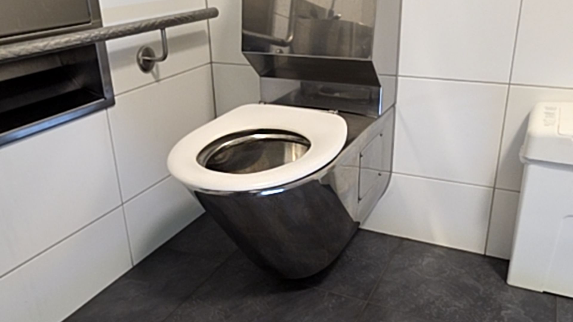 Stainless steel wall-hung toilet with white seat. Dark grey tiled floor, white tiled walls, and a textured horizontal stainless steel grab bar mounted on the left wall.