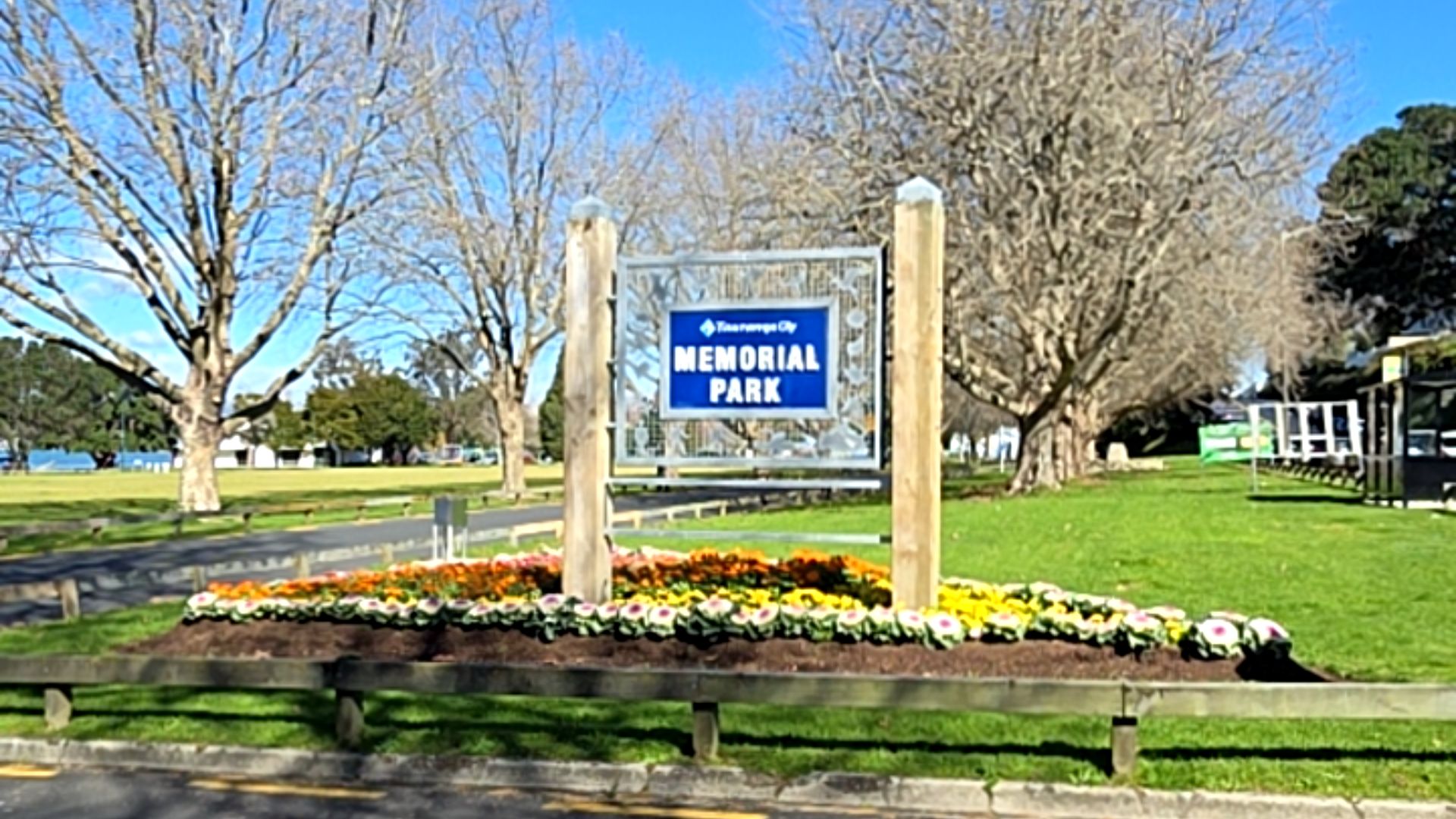 Tauranga City Memorial Park sign in blue between timber posts. Level asphalt pathway leads into the park. Low timber fence in foreground. Flat grassy area with bare trees.