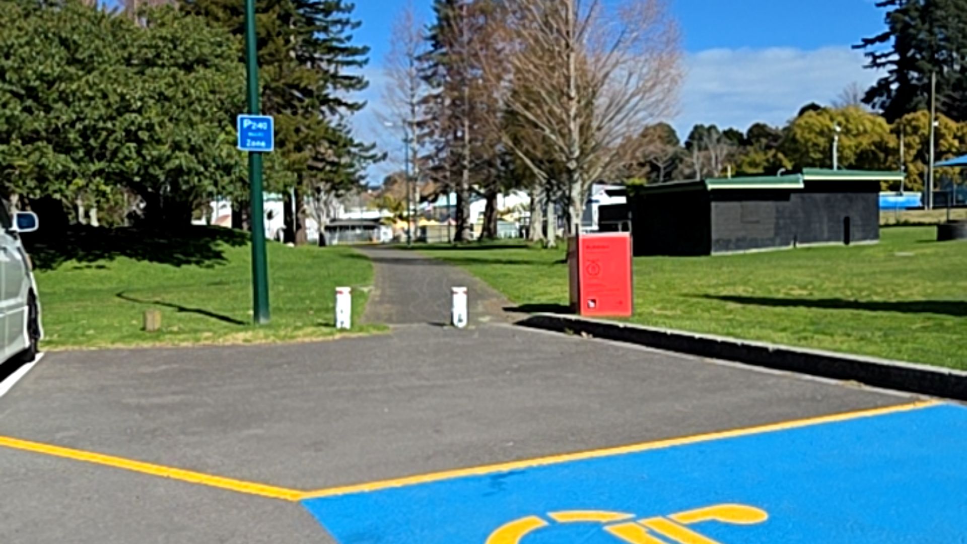 Level blue accessible parking space with yellow symbol. Wide, level asphalt pathway leads between two white bollards into a grassy park with trees and a blue parking sign.