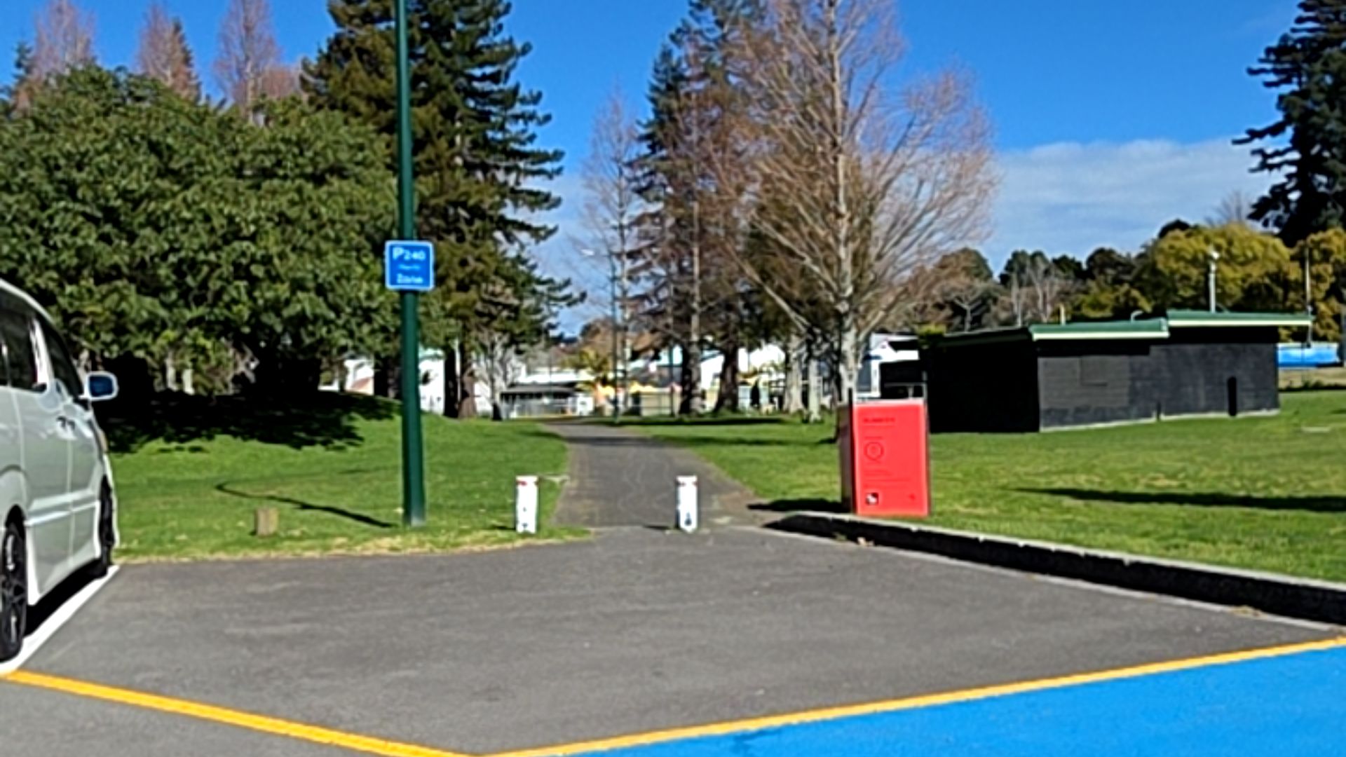Level asphalt pathway between white bollards leads from a car park with blue markings. Blue parking sign on left, red bin on right. Grassy parkland and mature trees in background.