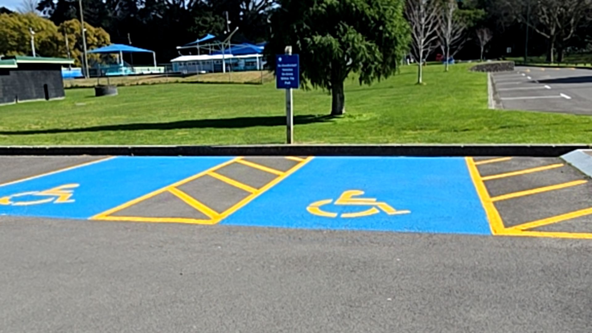 Two level blue-painted accessible parking bays with yellow wheelchair symbols and diagonal yellow hatched access aisles on asphalt, situated next to a grassed area with a blue sign.