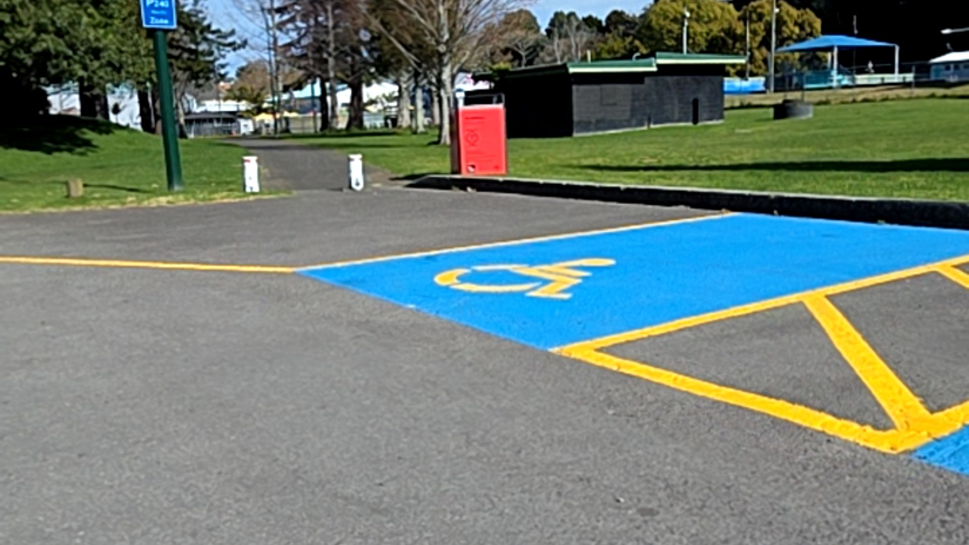 Level asphalt accessible parking space with blue surface and yellow wheelchair symbol. Yellow hatched buffer zone adjacent. A flat paved path leads into a grassy park area.