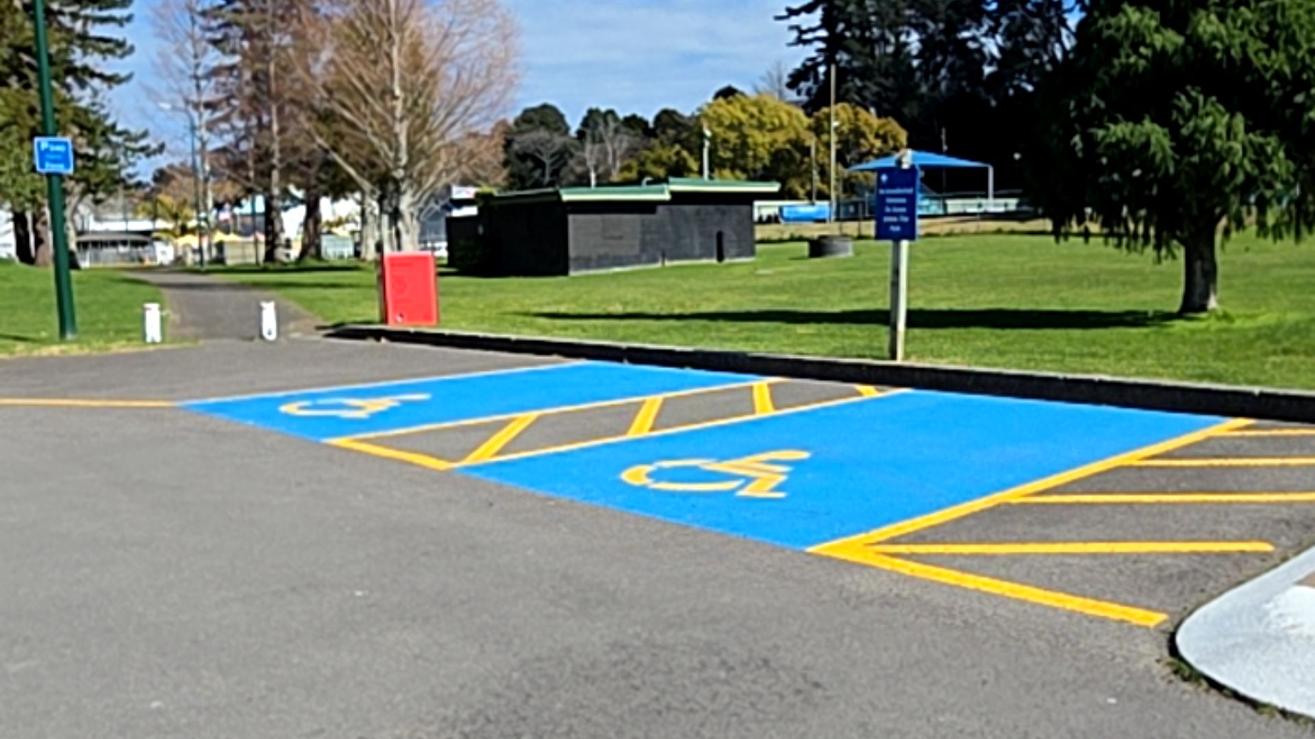 Two blue accessible parking bays with yellow wheelchair symbols and hatched access zones on level asphalt. A concrete kerb ramp is in the foreground with blue parking signage on a post nearby.