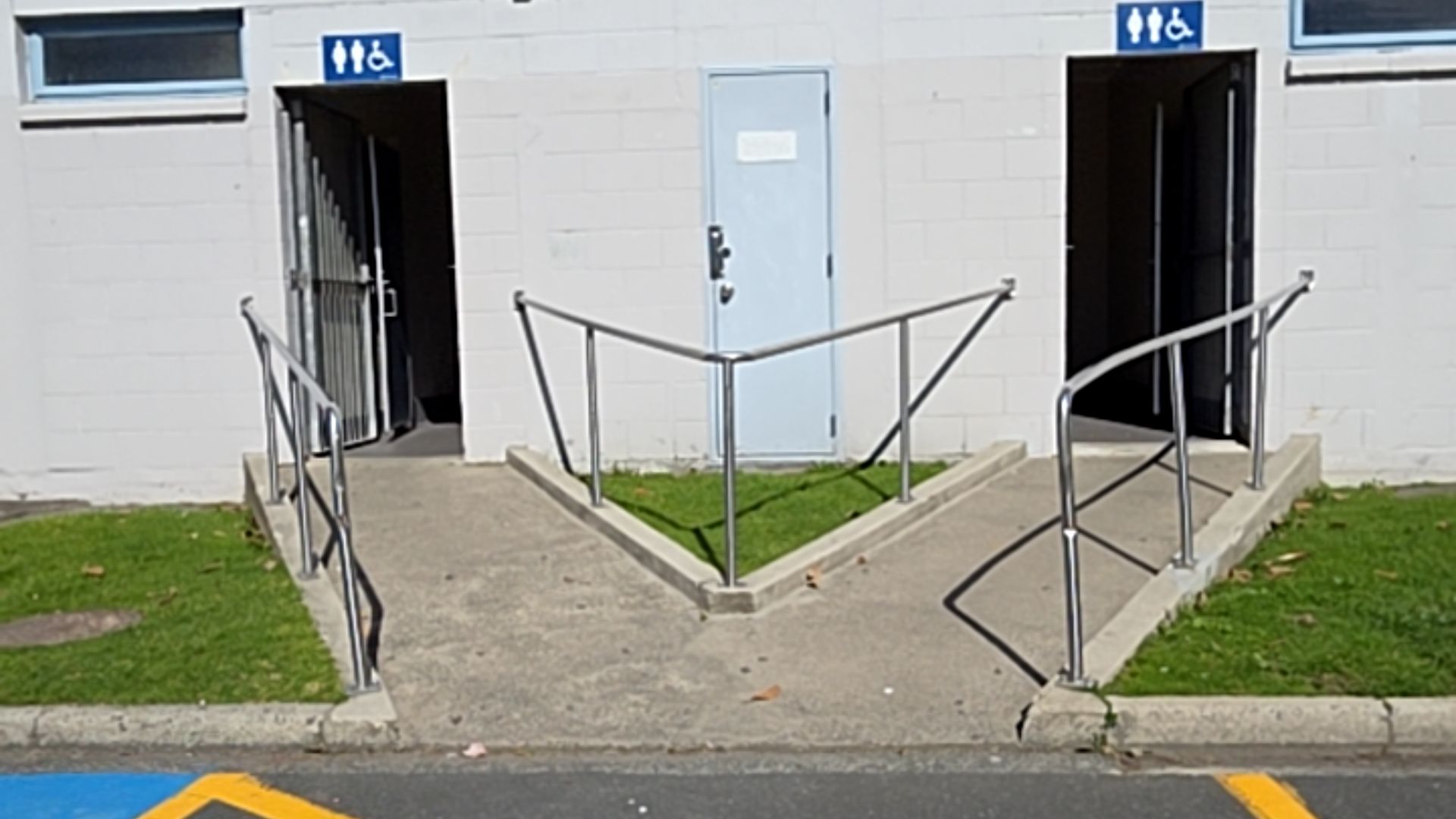 Two concrete ramps with metal handrails lead to accessible toilet entrances with blue signage. A central light blue door sits between the ramps. Yellow and blue markings on bitumen foreground.