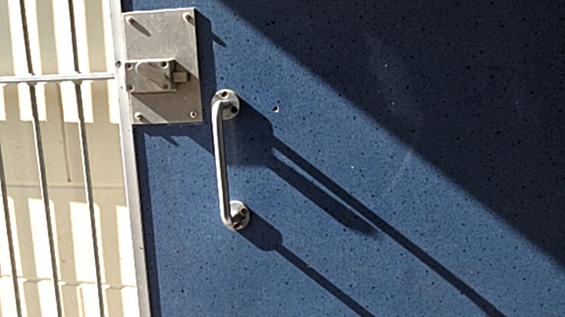 Blue door with vertical silver-coloured pull handle and surface-mounted metal latch. Cream-coloured slatted fence to the left. Sunlight casts long shadows across the door surface.