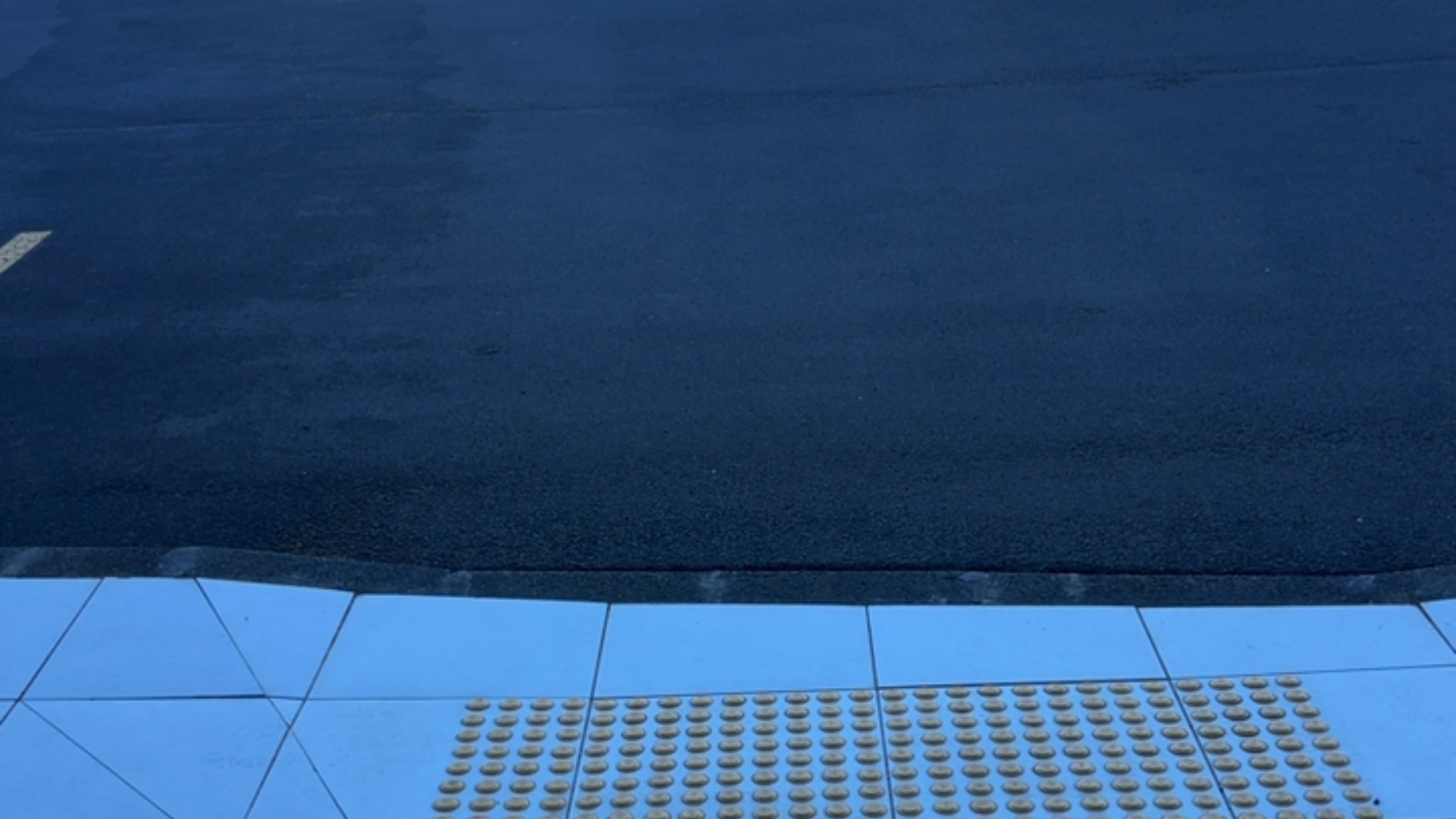 Light blue tiled pavement with yellow tactile studs, leading to a level transition across a dark grey kerb edge to a bitumen road surface.