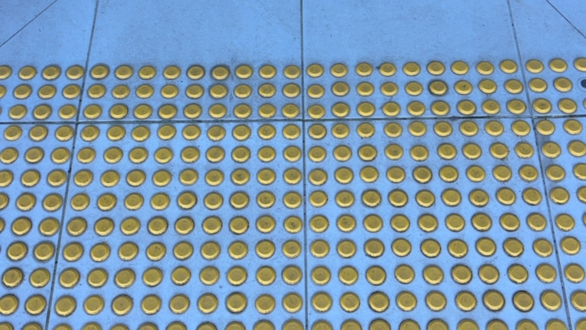Light blue tiled floor surface with a grid pattern of raised yellow circular tactile studs. Thin grout lines divide the floor into large square sections.