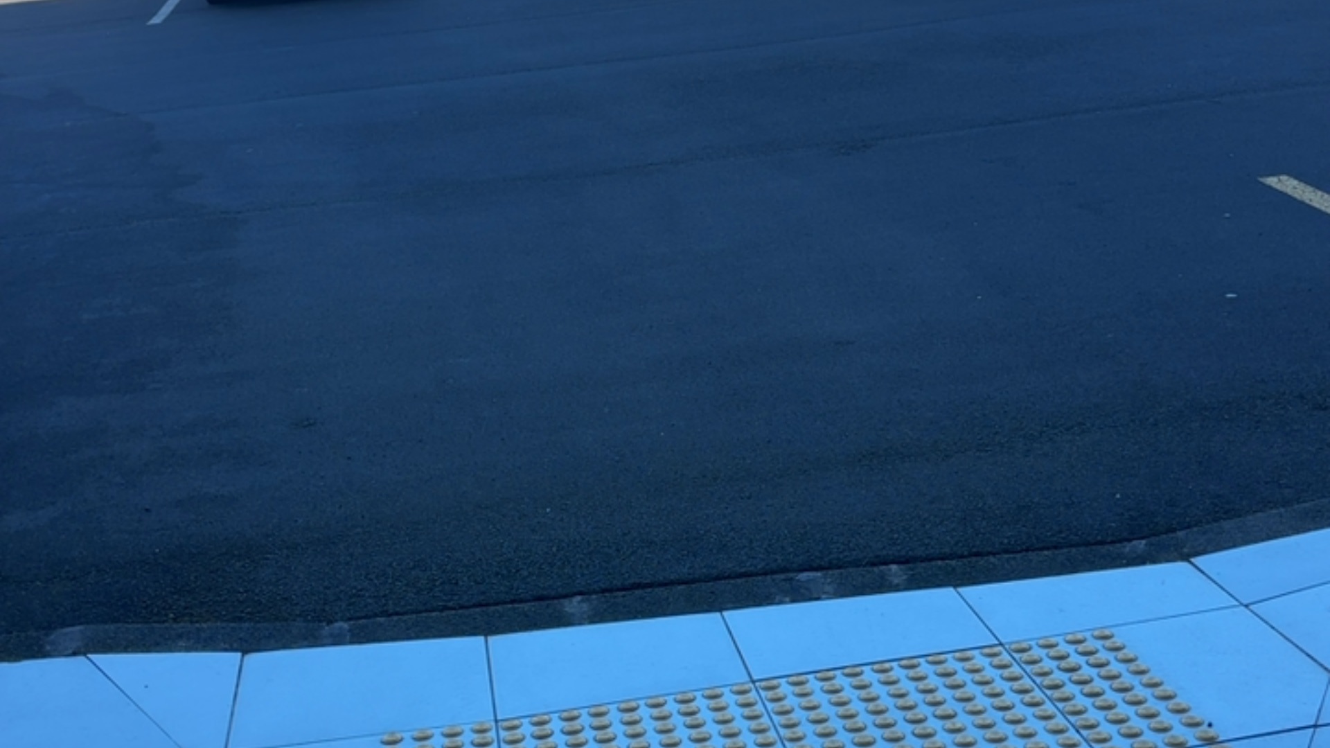 Light-coloured tiled pavement featuring yellow tactile ground surface indicators at a level kerb transition to a dark asphalt road.