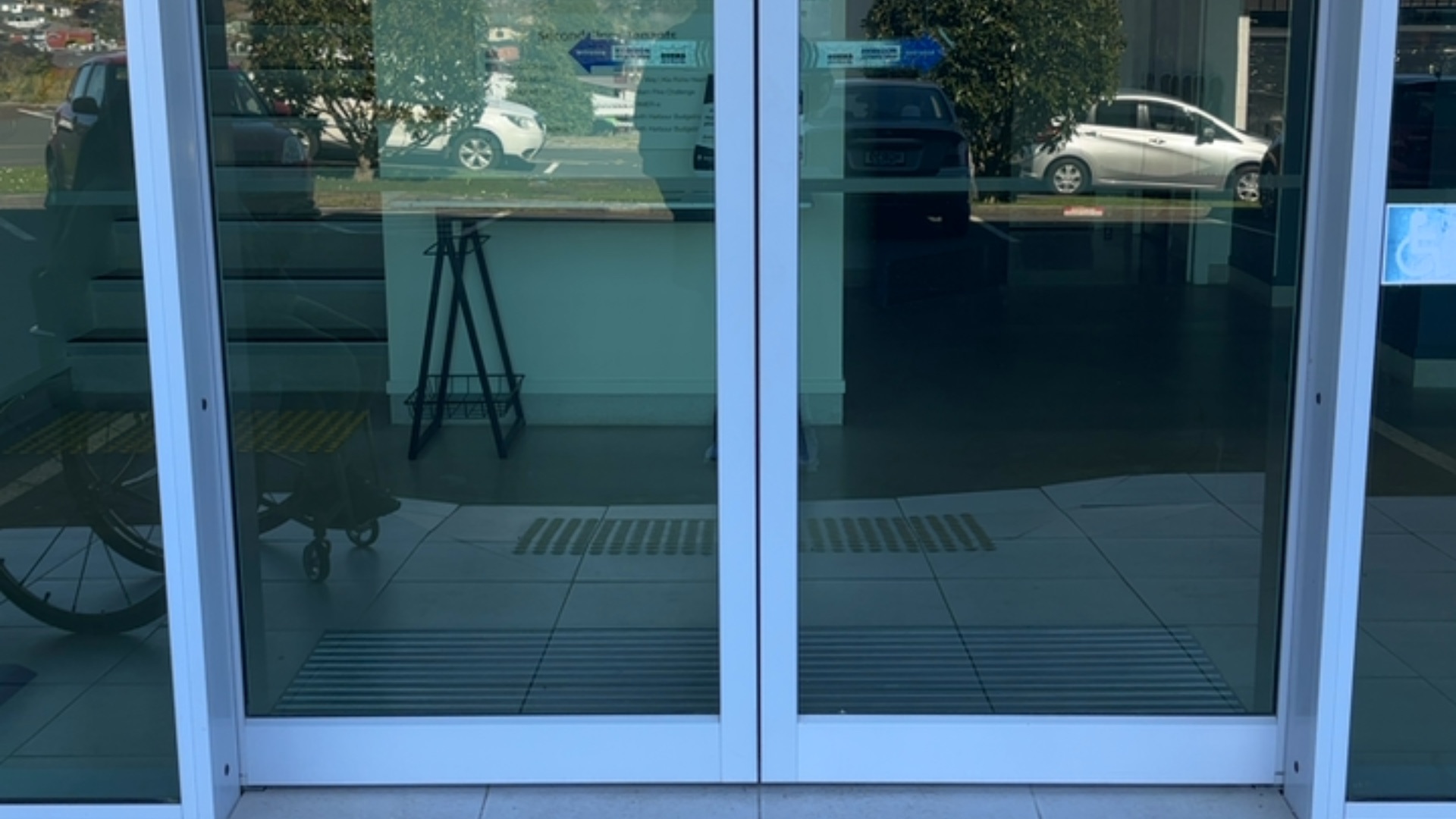 Double automatic glass sliding doors with white frames and level entry on tiled floor. Tactile paving strips on the interior floor. Small blue wheelchair symbol sticker on the right door.