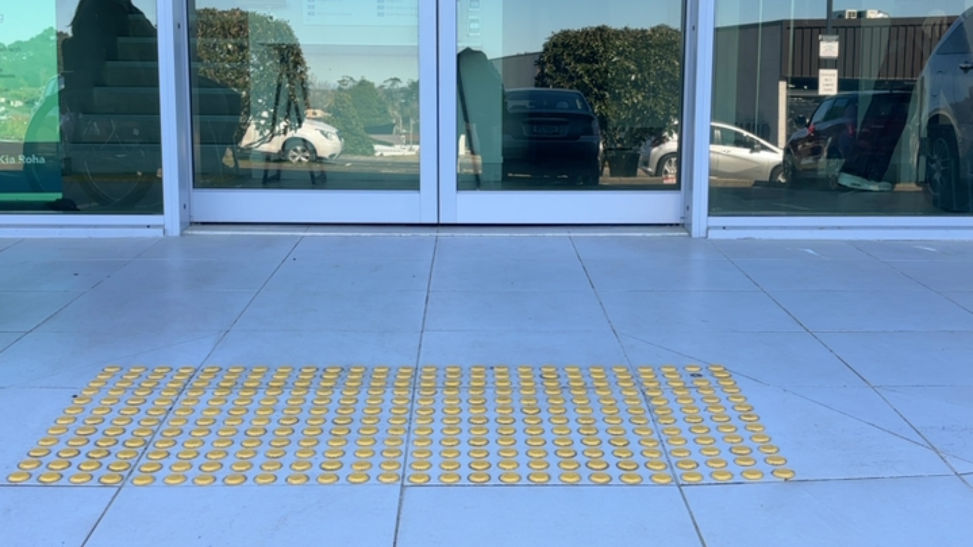 Level entrance with glass doors. The light-coloured tiled floor features a rectangular patch of yellow raised tactile studs located directly in front of the doorway.
