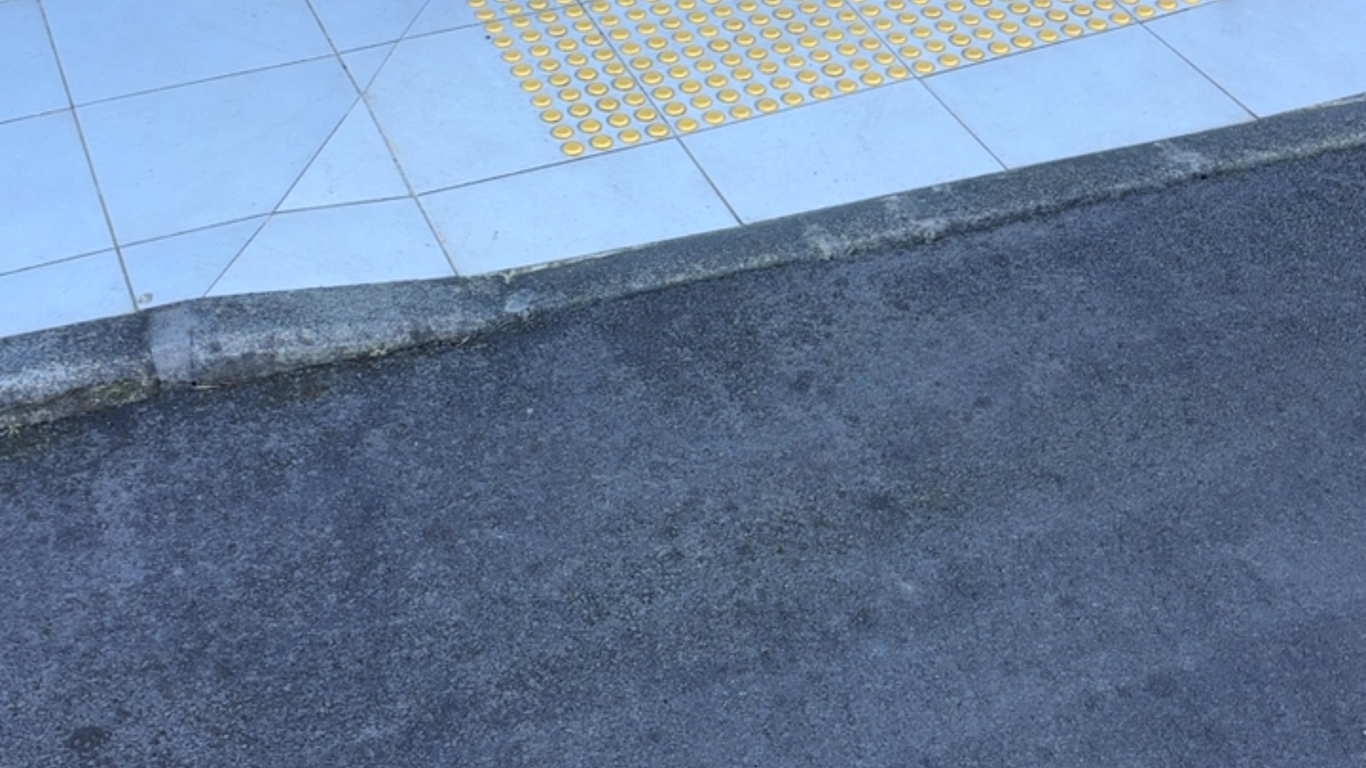 Asphalt pathway leads to a stone kerb and light blue tiled area with a section of bright yellow tactile ground surface indicators featuring raised circular studs.