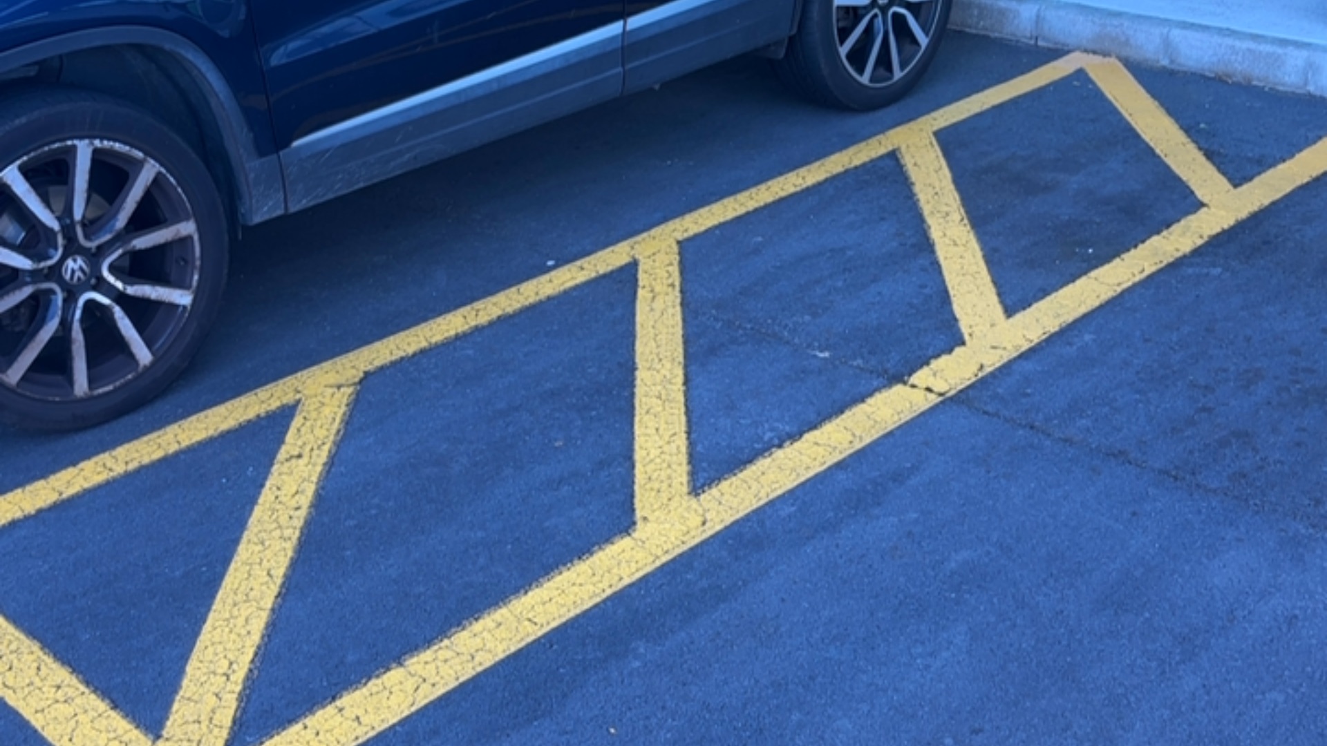 Yellow cross-hatched access aisle on level dark asphalt, located beside a parked dark blue car and a concrete kerb.