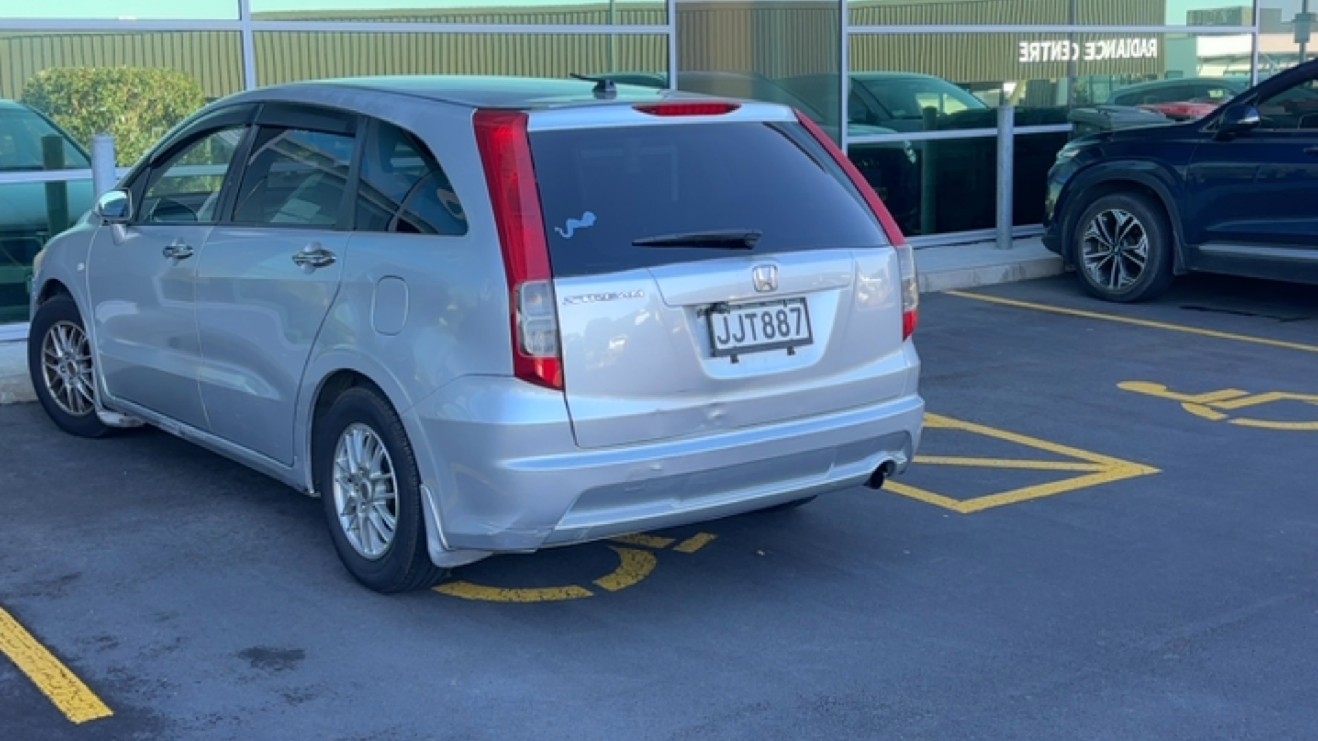 Silver car parked in a designated accessible parking space marked with yellow wheelchair symbols and hatch markings on a level bitumen surface.
